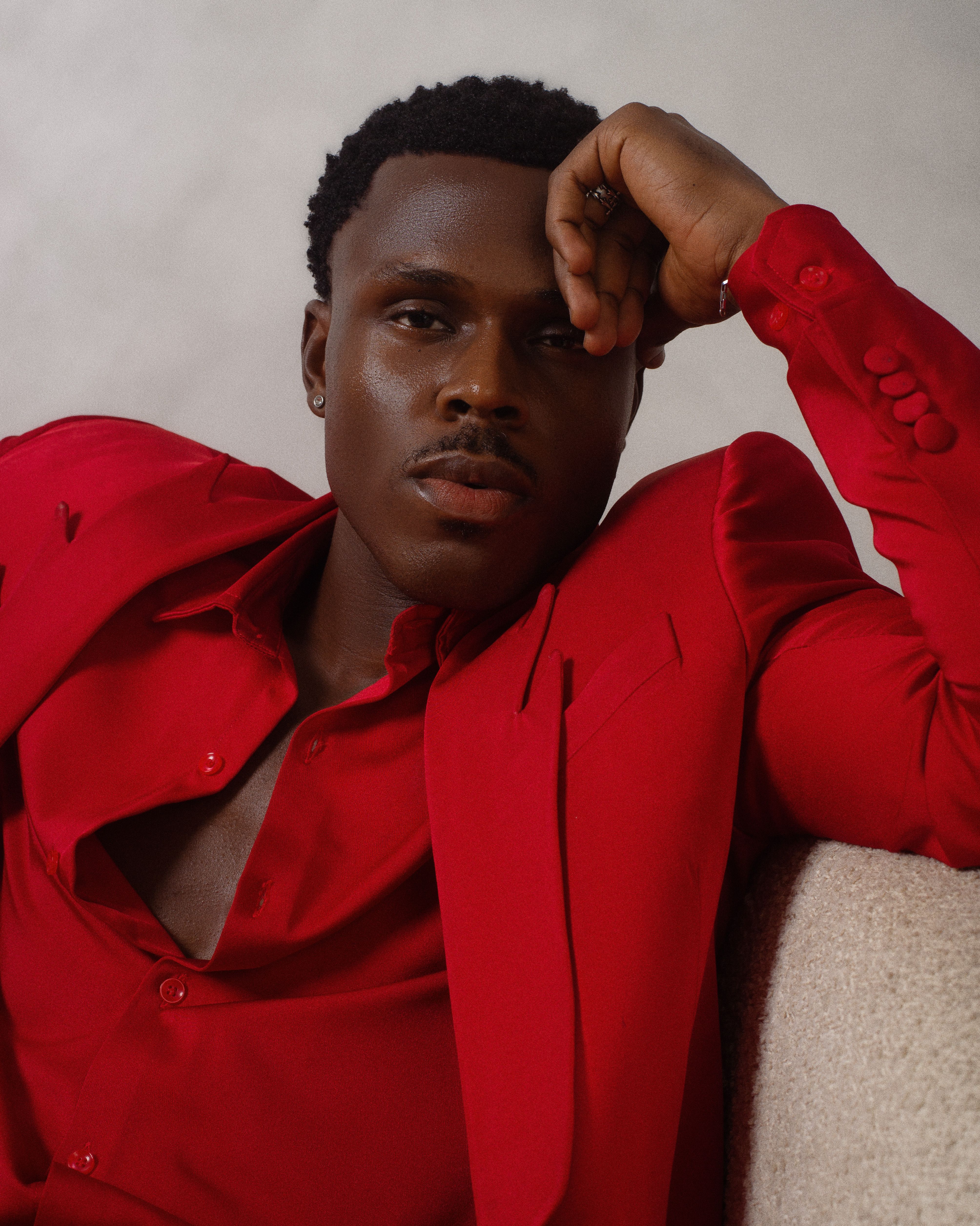 A stylish man wearing a red suit relaxes indoors, exuding confidence and elegance.