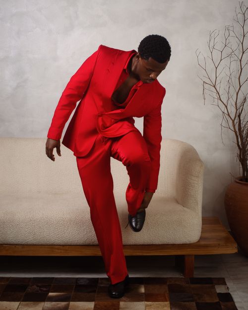 Free Man in a red suit poses confidently on a beige sofa against a neutral background. Stock Photo