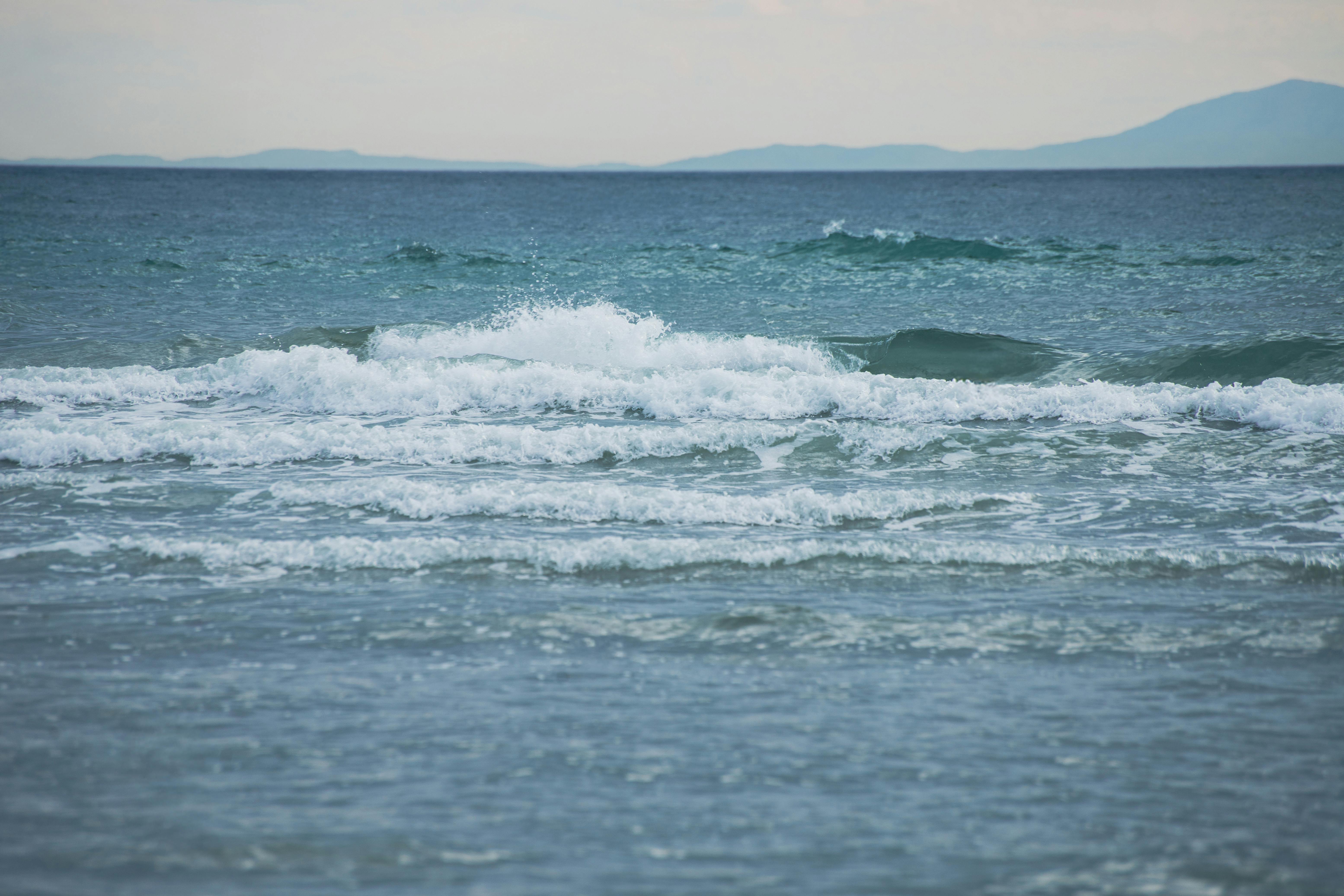 Δωρεάν Ήρεμα κύματα του ωκεανού σκάνε στην ακτή με μακρινούς λόφους να φαίνονται κάτω από έναν γαλήνιο ουρανό. Φωτογραφία από στοκ φωτογραφιών