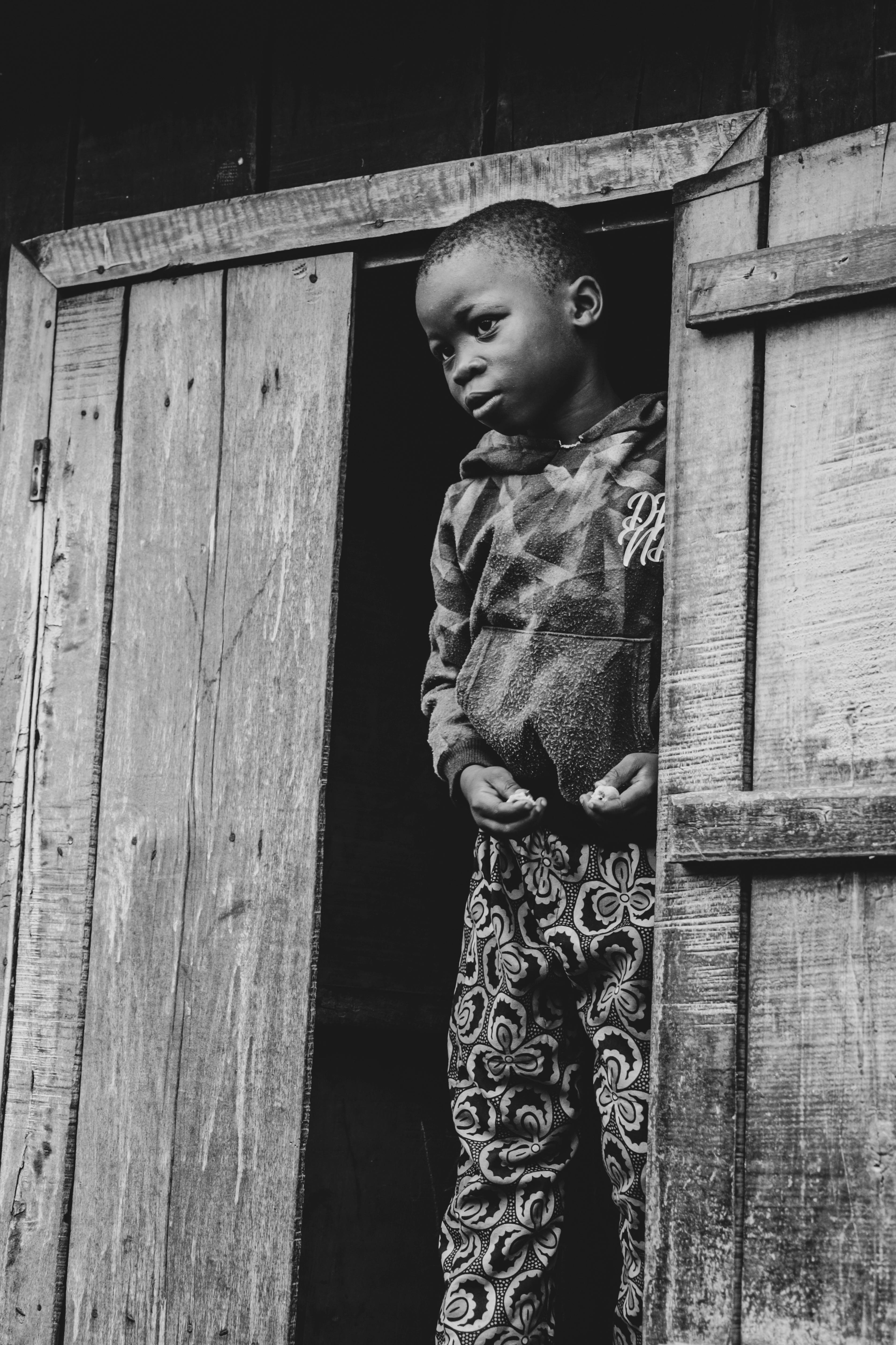 Kostenlos Ein nachdenkliches Kind steht in einem hölzernen Türrahmen, eingefangen in Schwarzweiß. Stock-Foto