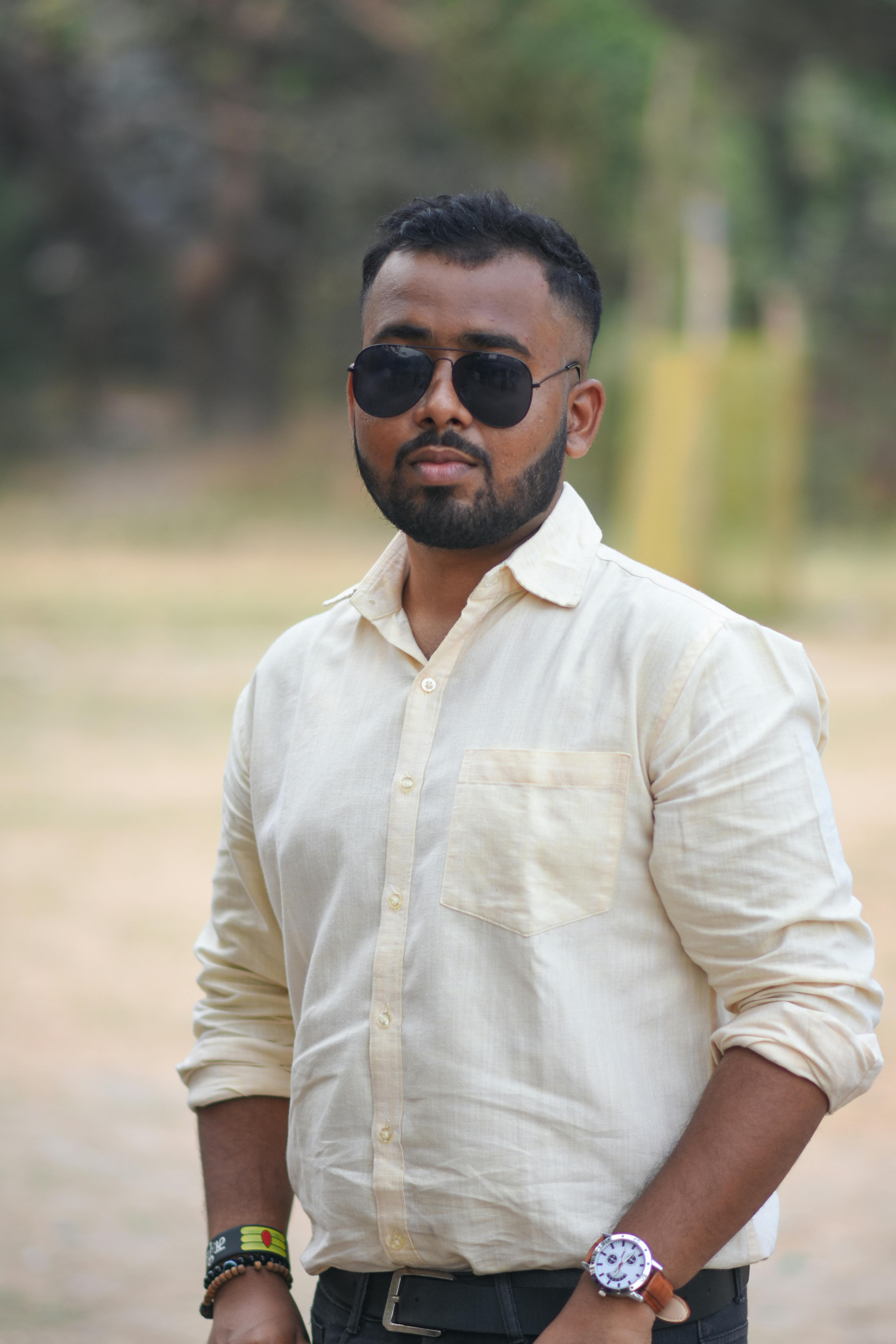 A man in sunglasses and a beige shirt poses confidently outdoors.