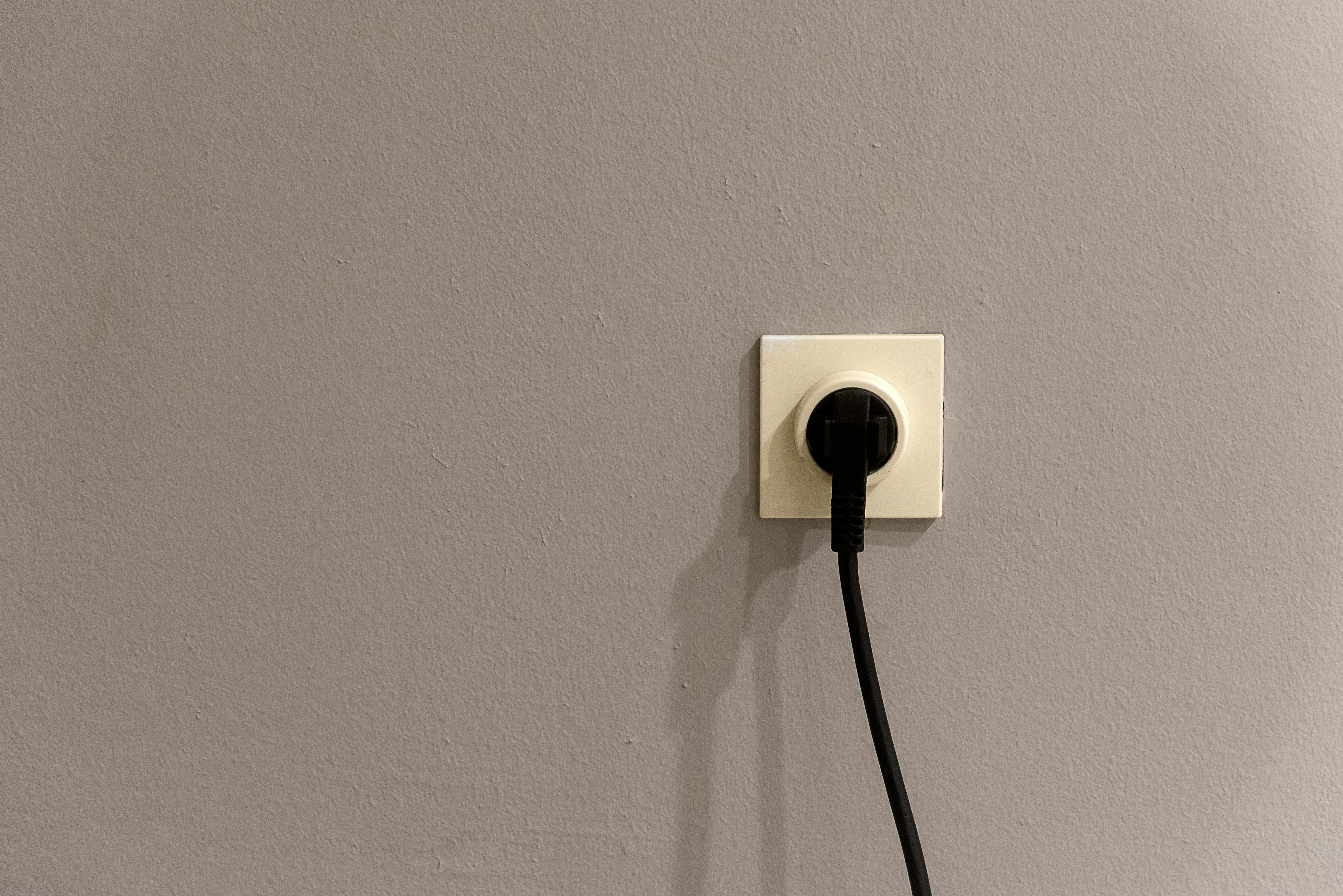 A black plug inserted into a white wall socket on a plain grey wall.