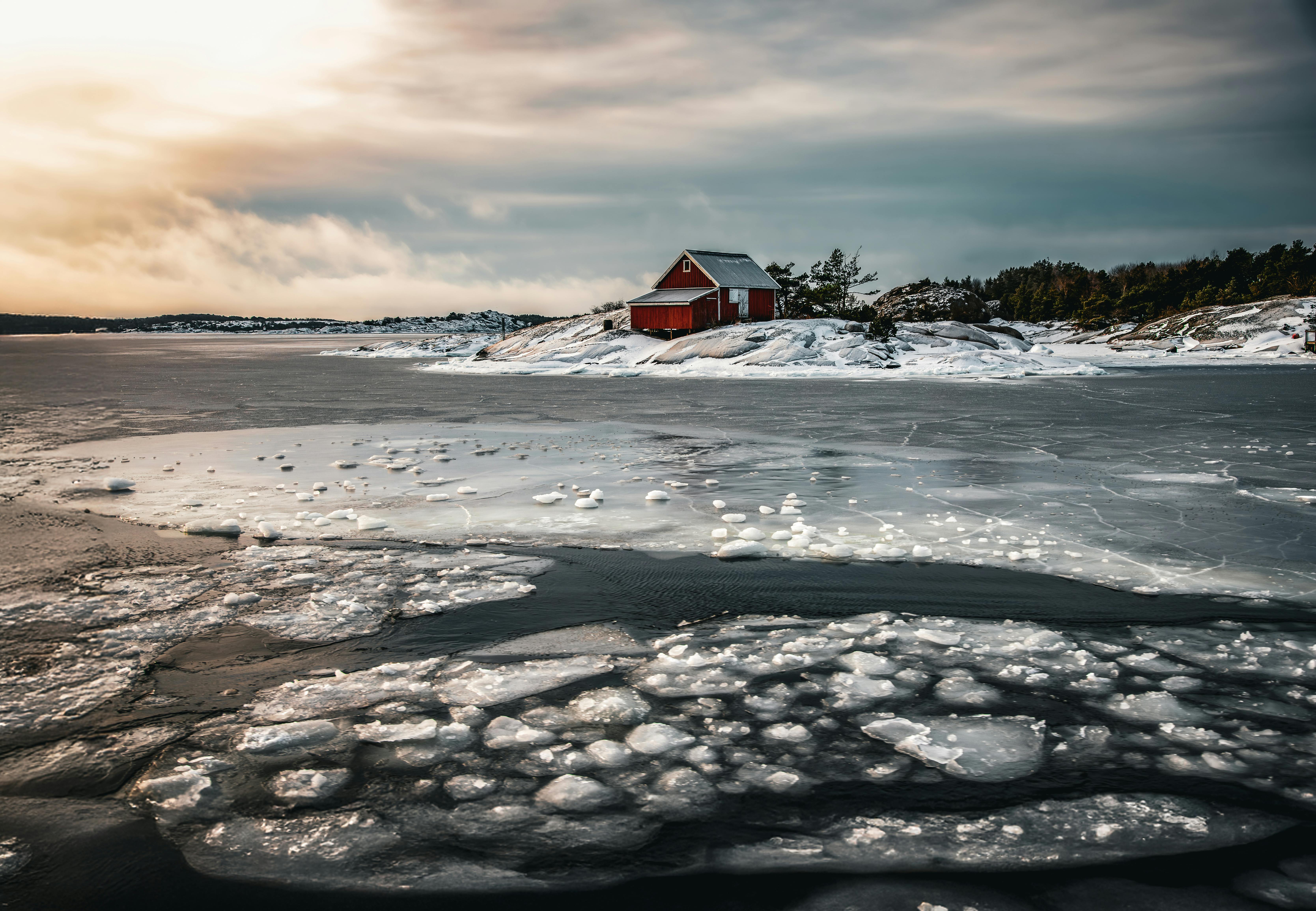 Kostenlos Idyllische Winterszene an einer eisigen Küste mit einer einsamen roten Hütte im Sonnenuntergang. Stock-Foto