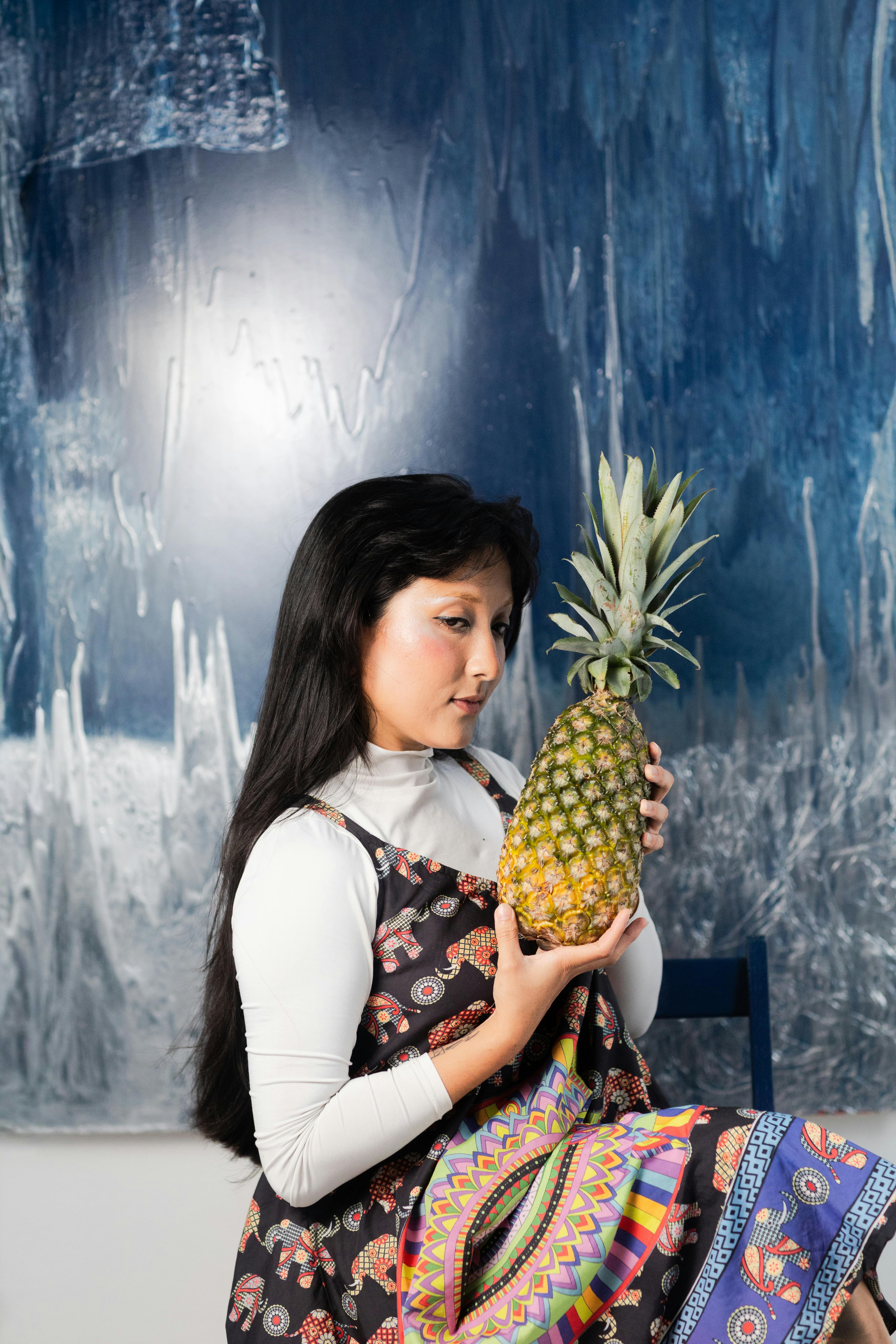 Gratis En kvinne ser på en ananas foran et abstrakt blått kunstverk. Arkivbilde