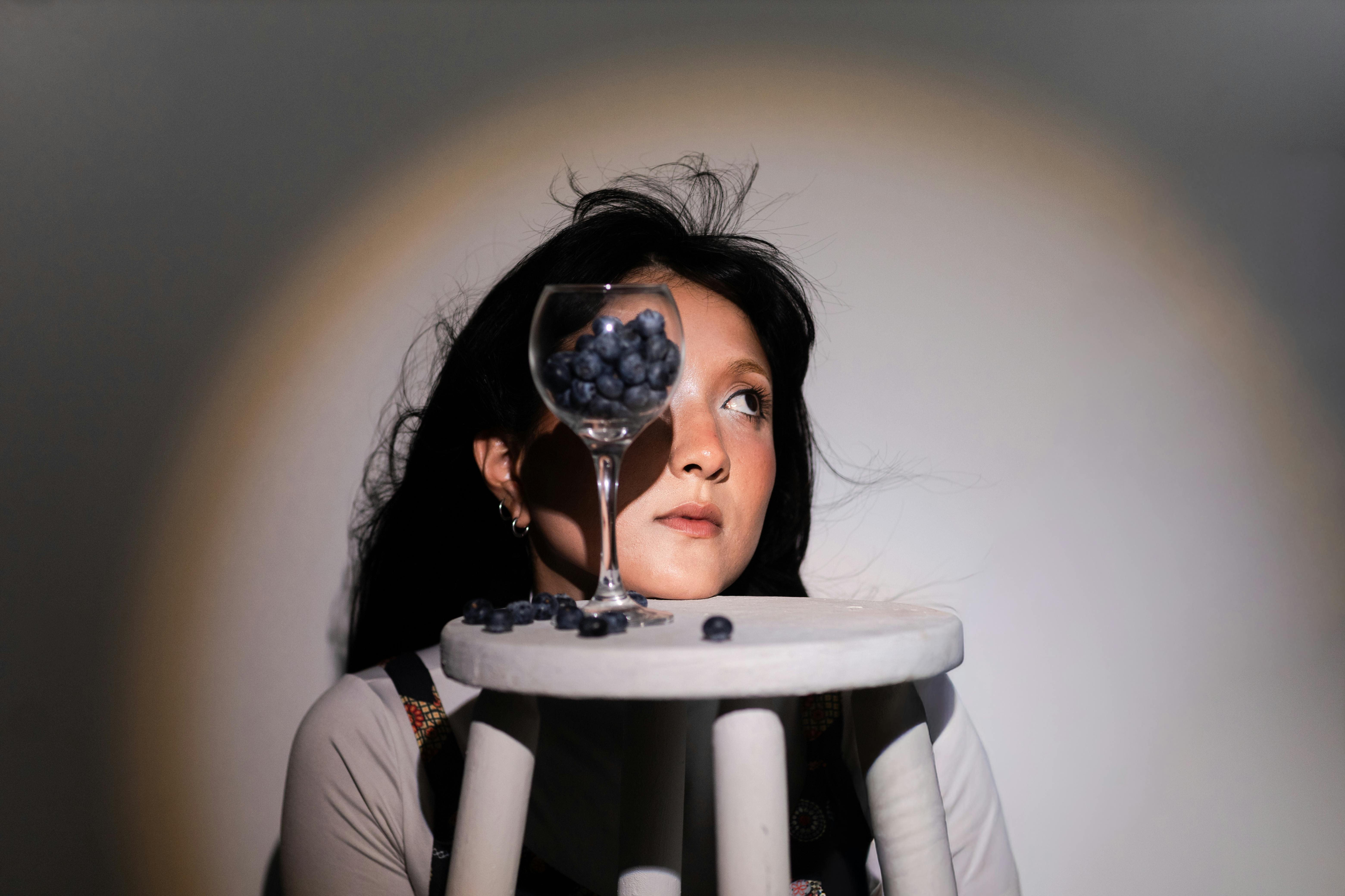 Creative portrait featuring a young woman with a glass of blueberries on a stool.