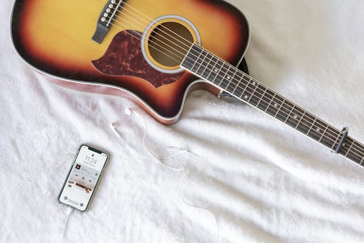 Acoustic guitar with smartphone playing music, headphones on soft white fabric.