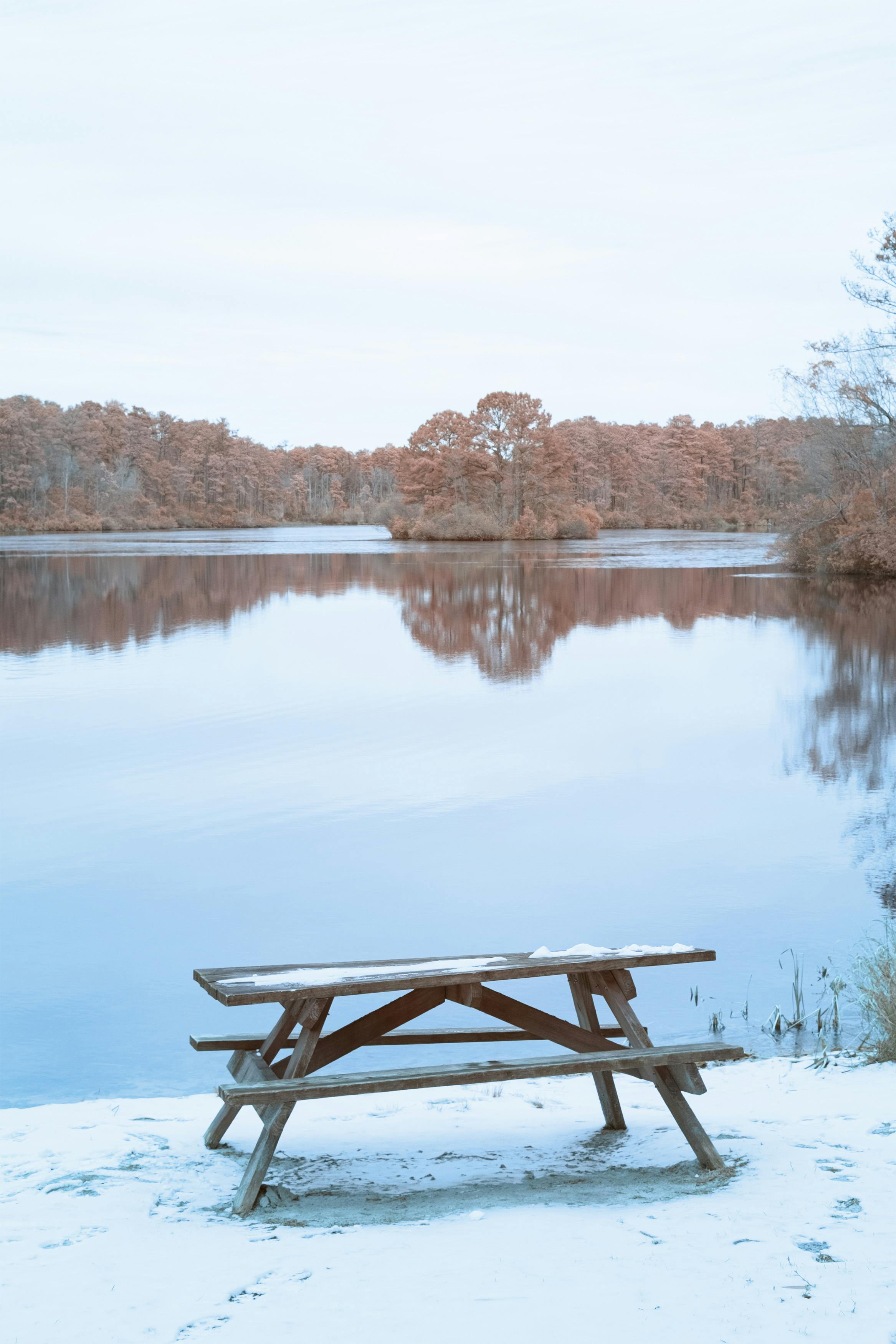 A serene winter landscape featuring a snow-covered bench by a peaceful, reflective lake.