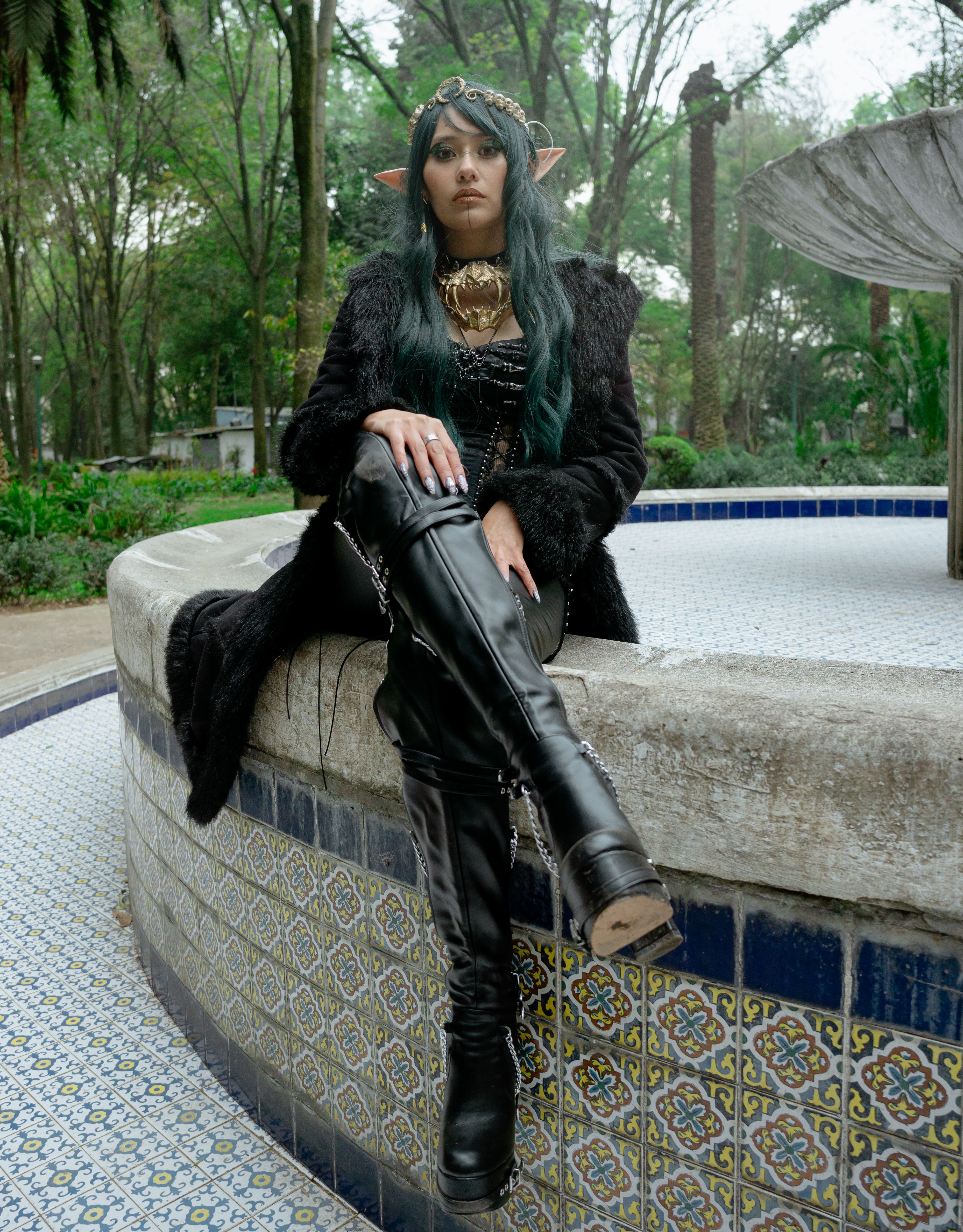 A woman dressed as a fantasy elf sits by a decorative fountain in an outdoor park.