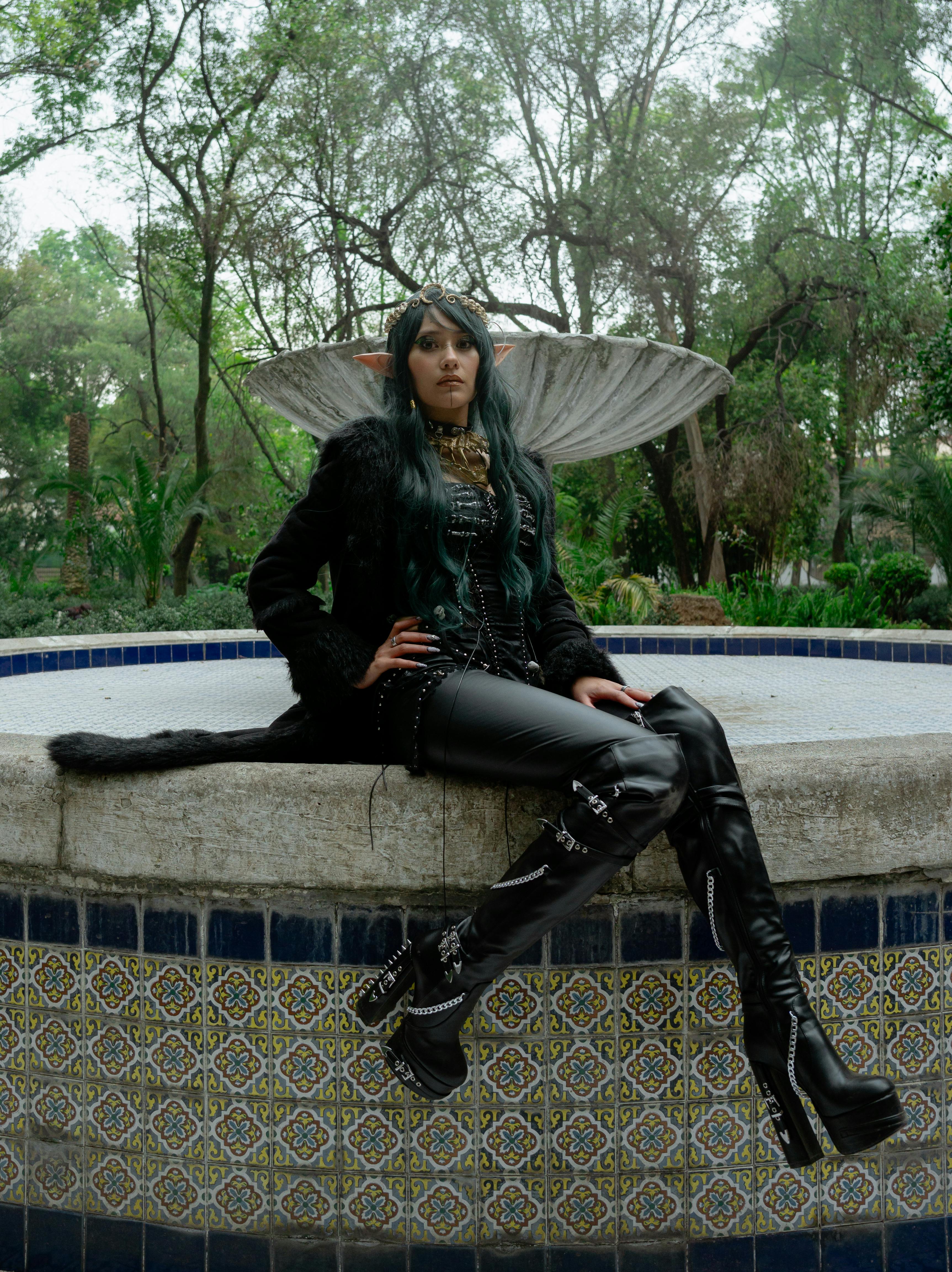 A striking dark fairy in black attire sits confidently outdoors in a mystical forest.