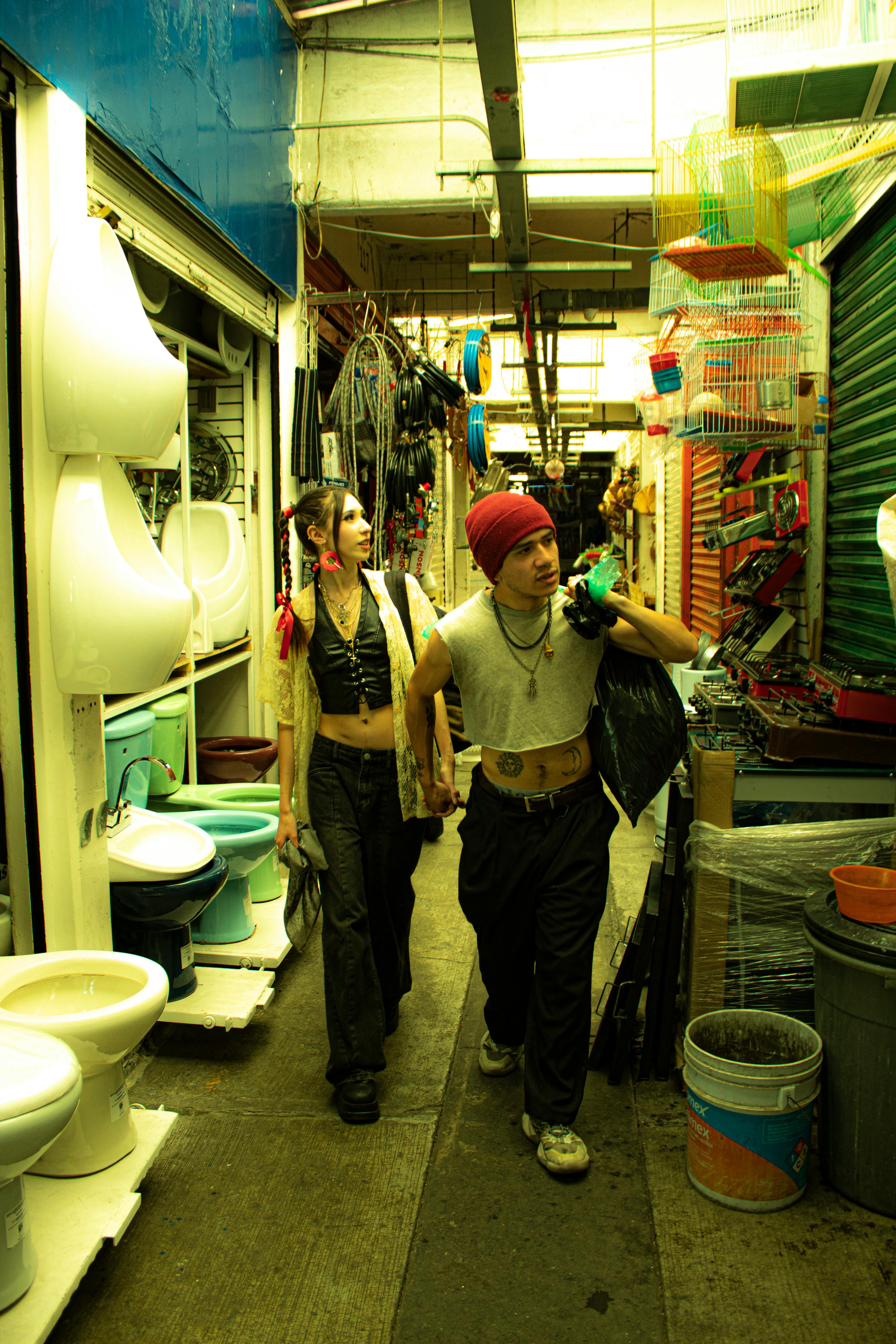 Gratis To unge voksne går gjennom en livlig markedsgang fylt med diverse varer og sterk belysning. Arkivbilde