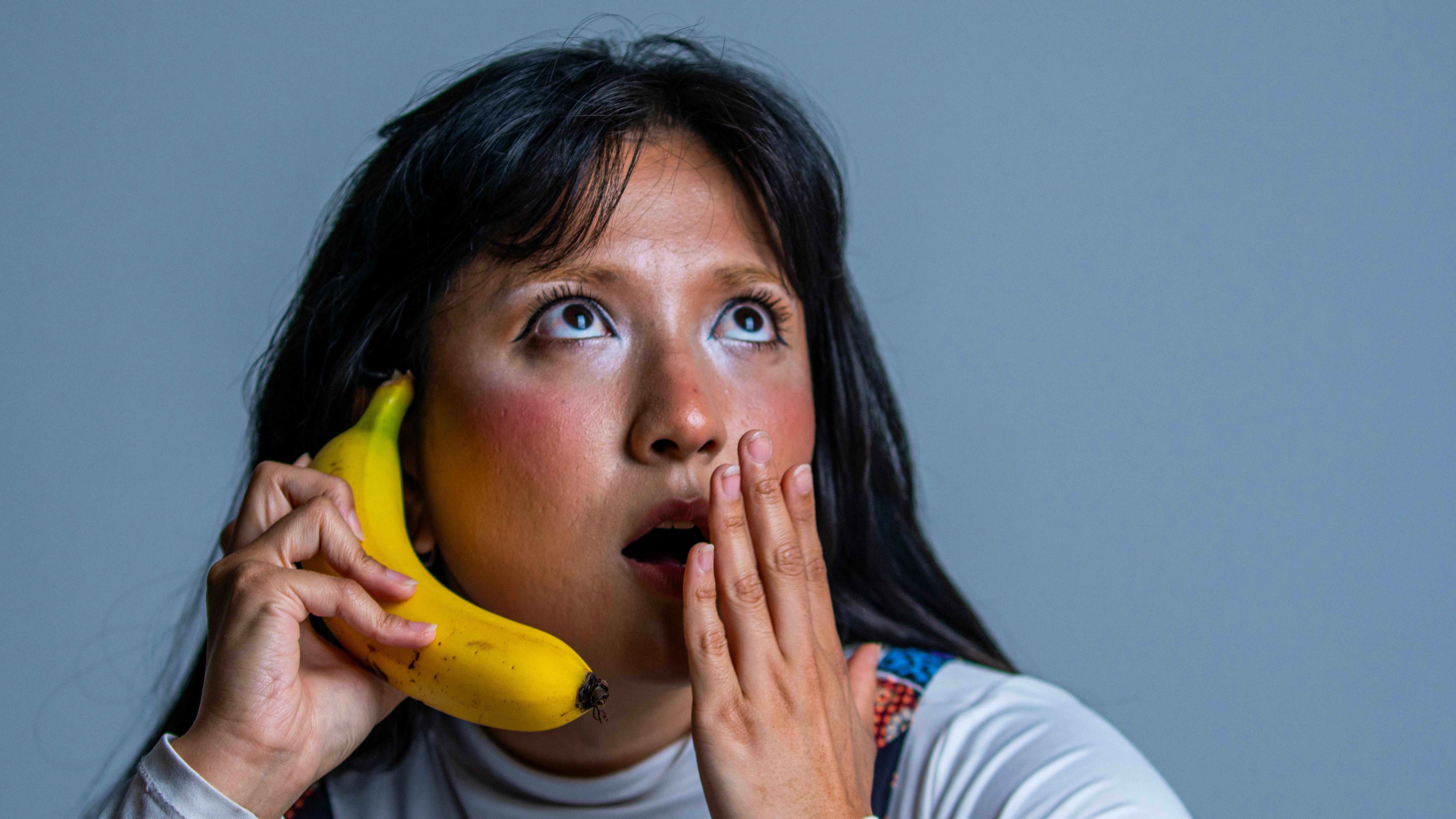 Free A surprised woman holding a banana like a phone against a neutral background. Stock Photo