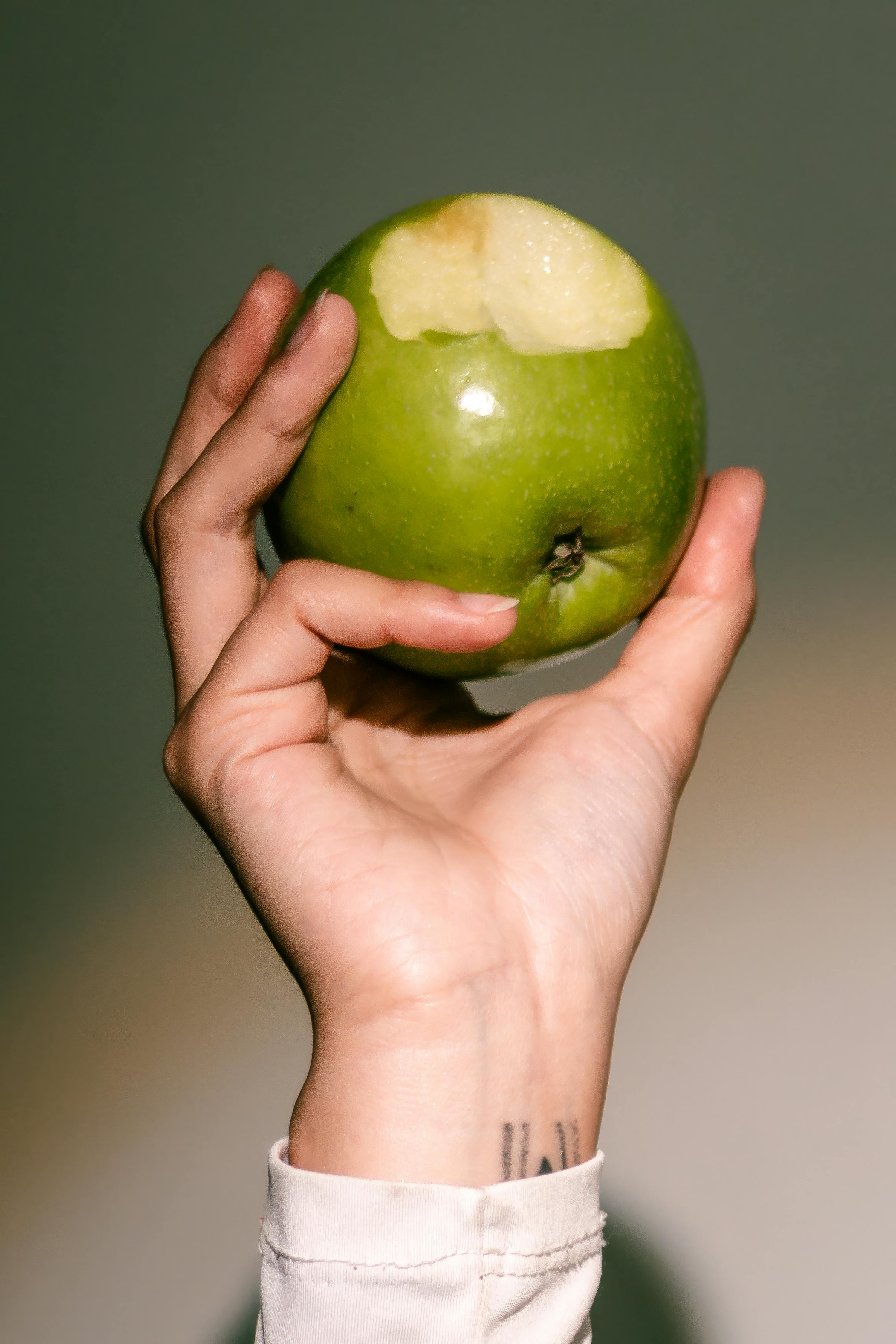 Free A hand holding a bitten green apple, captured indoors with natural light and a muted background. Stock Photo