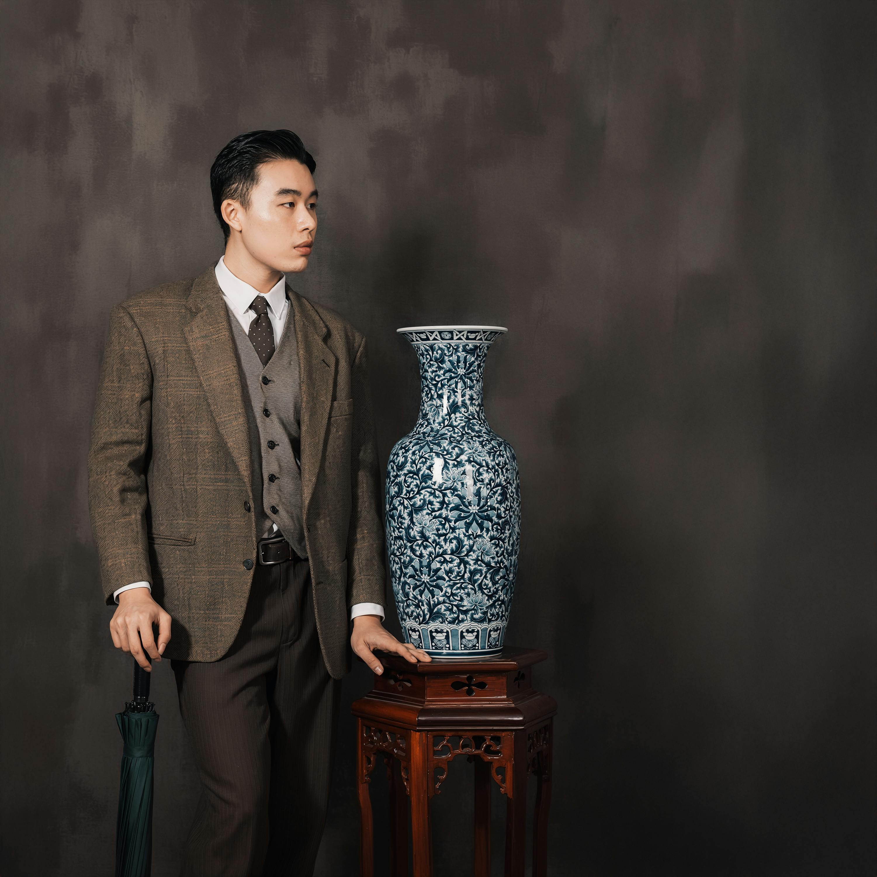 Portrait of a man in vintage attire standing next to a neo-classical blue ceramic vase on a wooden table.