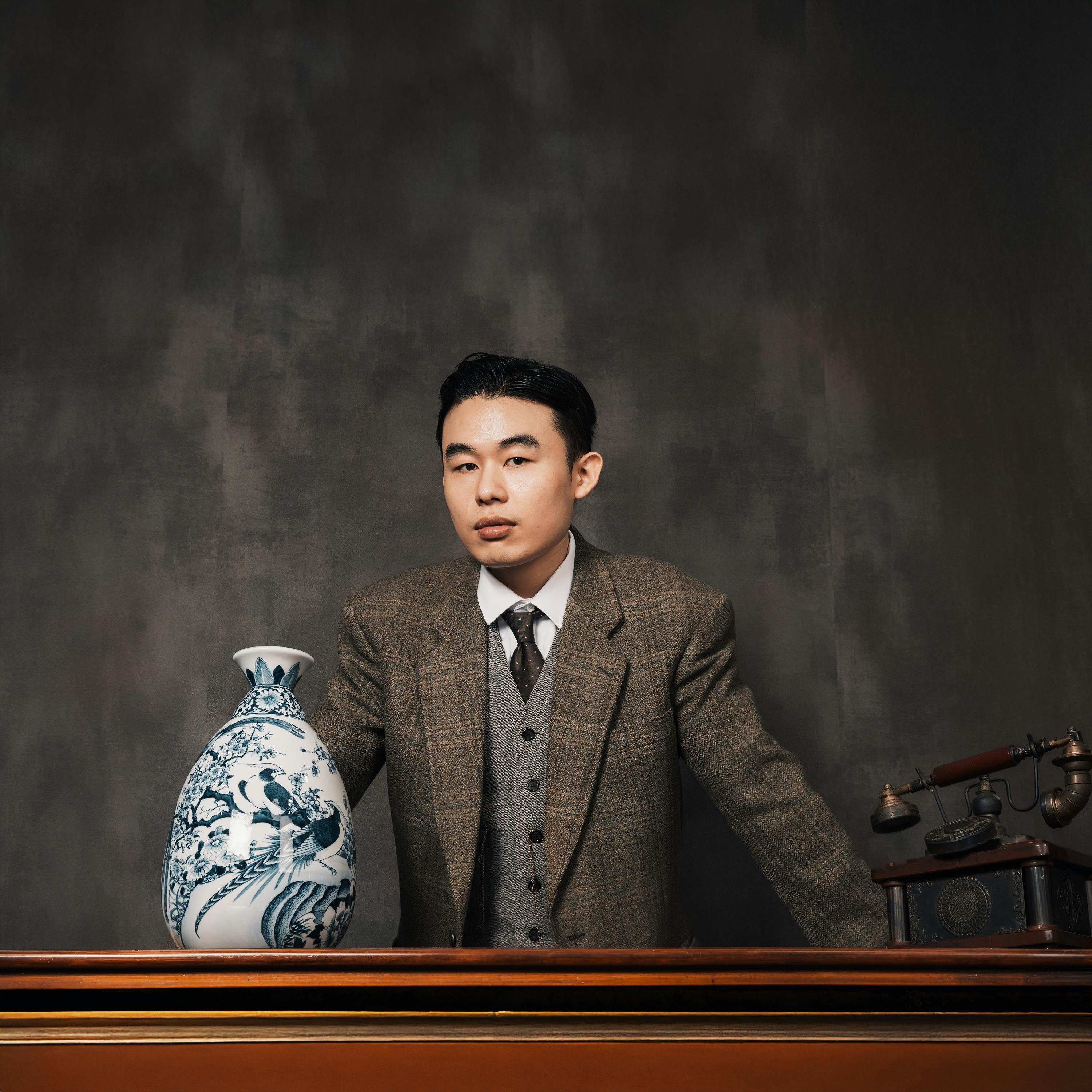 A portrait of a man in a suit, standing beside an elegant ceramic vase indoors.