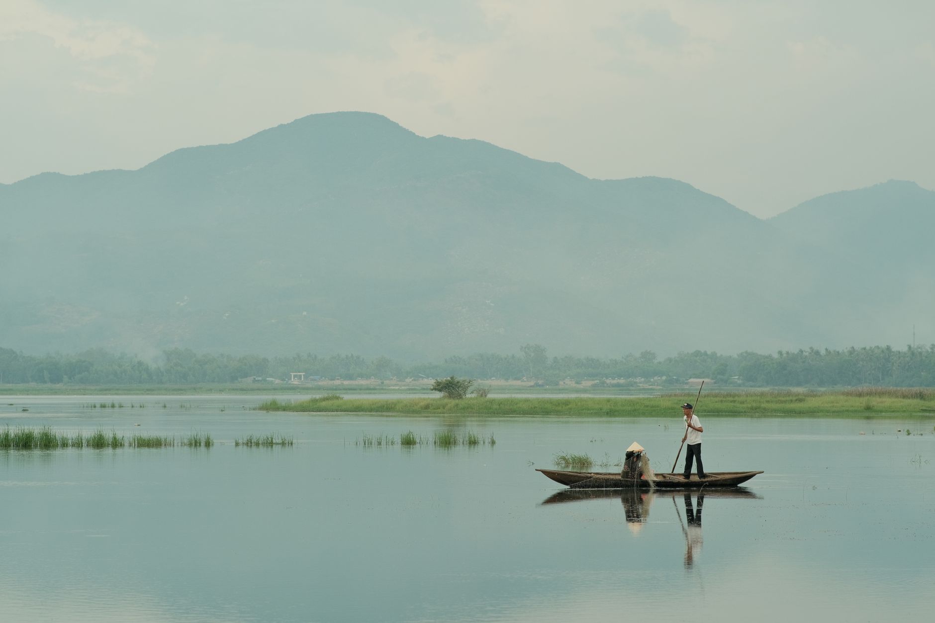Two fishermen on a boat in a serene Vietnamese lake against a mountainous backdrop.