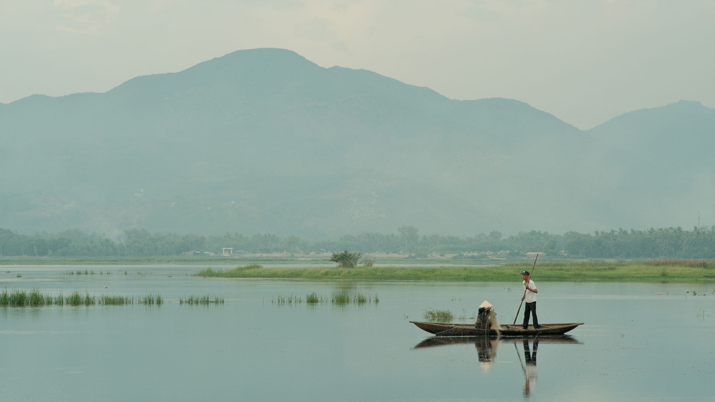 Two fishermen on a boat in a serene Vietnamese lake against a mountainous backdrop.