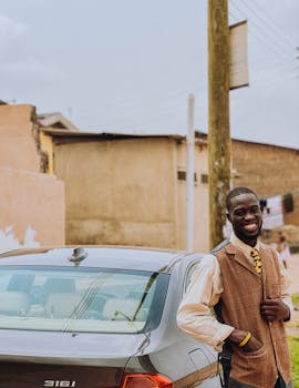 A cheerful man stands beside a car on a street in Accra, Ghana, exuding a joyful and relaxed vibe.