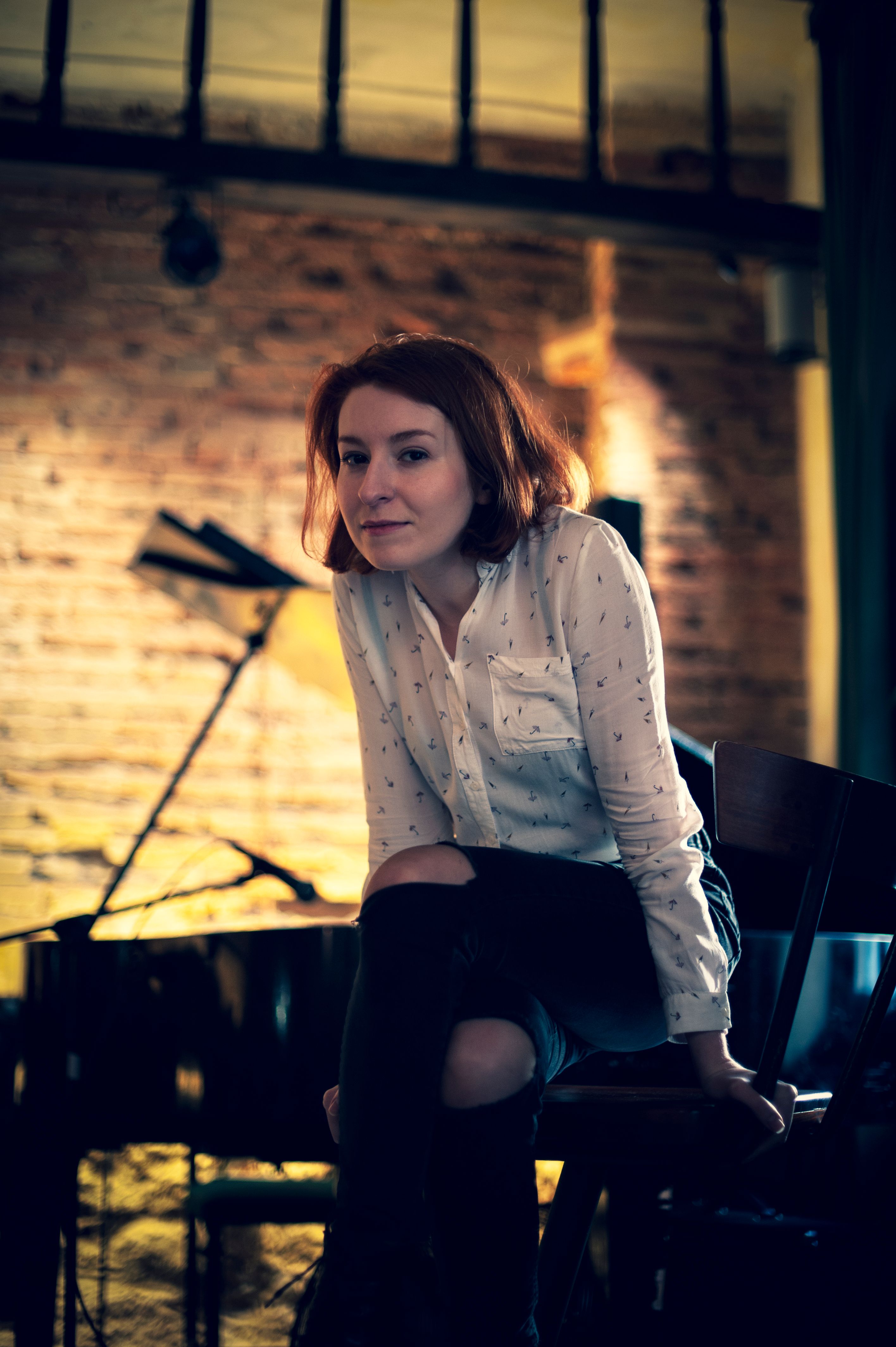 A thoughtful adult woman in a white blouse sits near a grand piano in an atmospheric indoor setting.