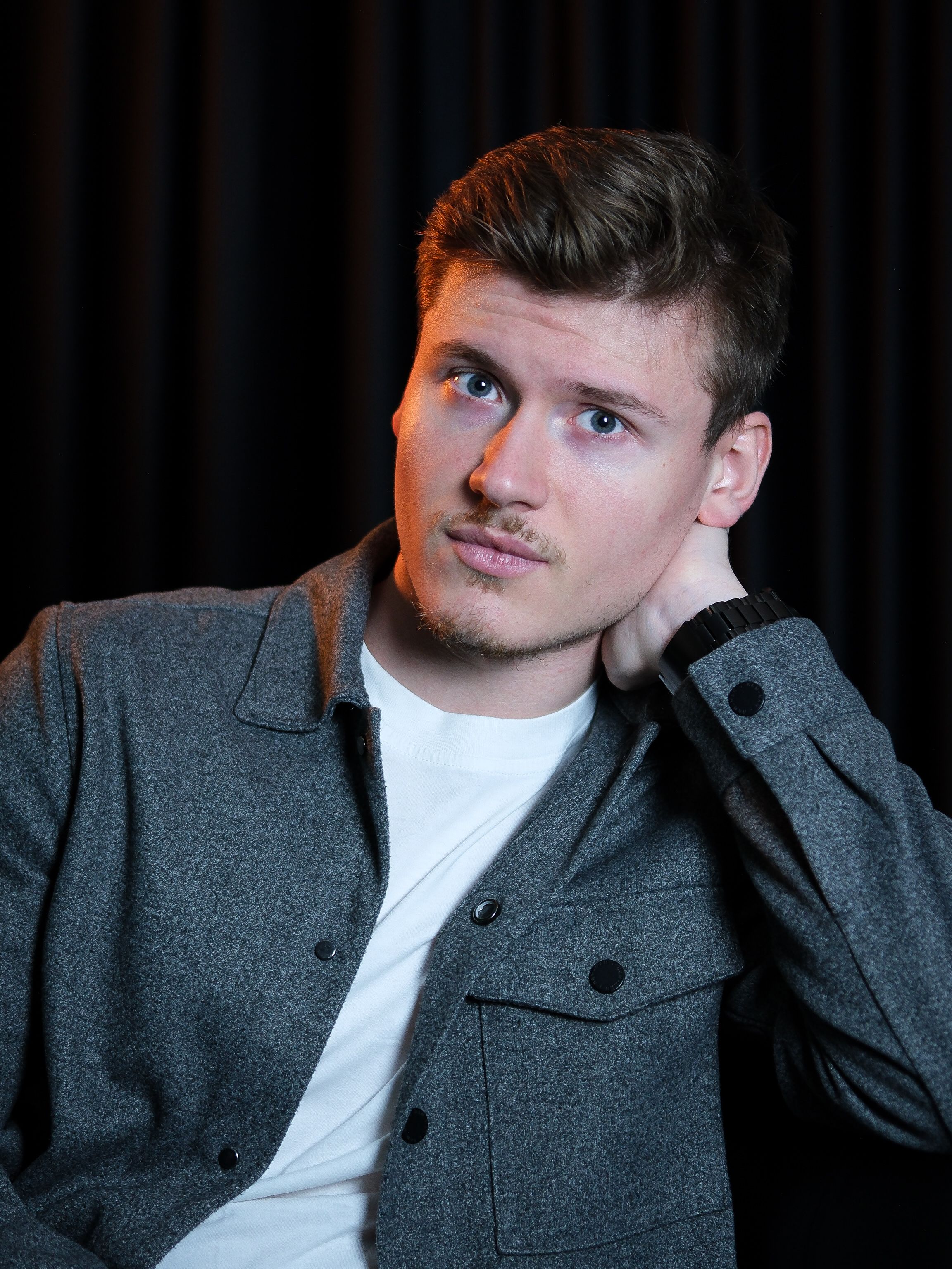 Portrait of a young adult man in a gray jacket against a dark background, looking thoughtful.