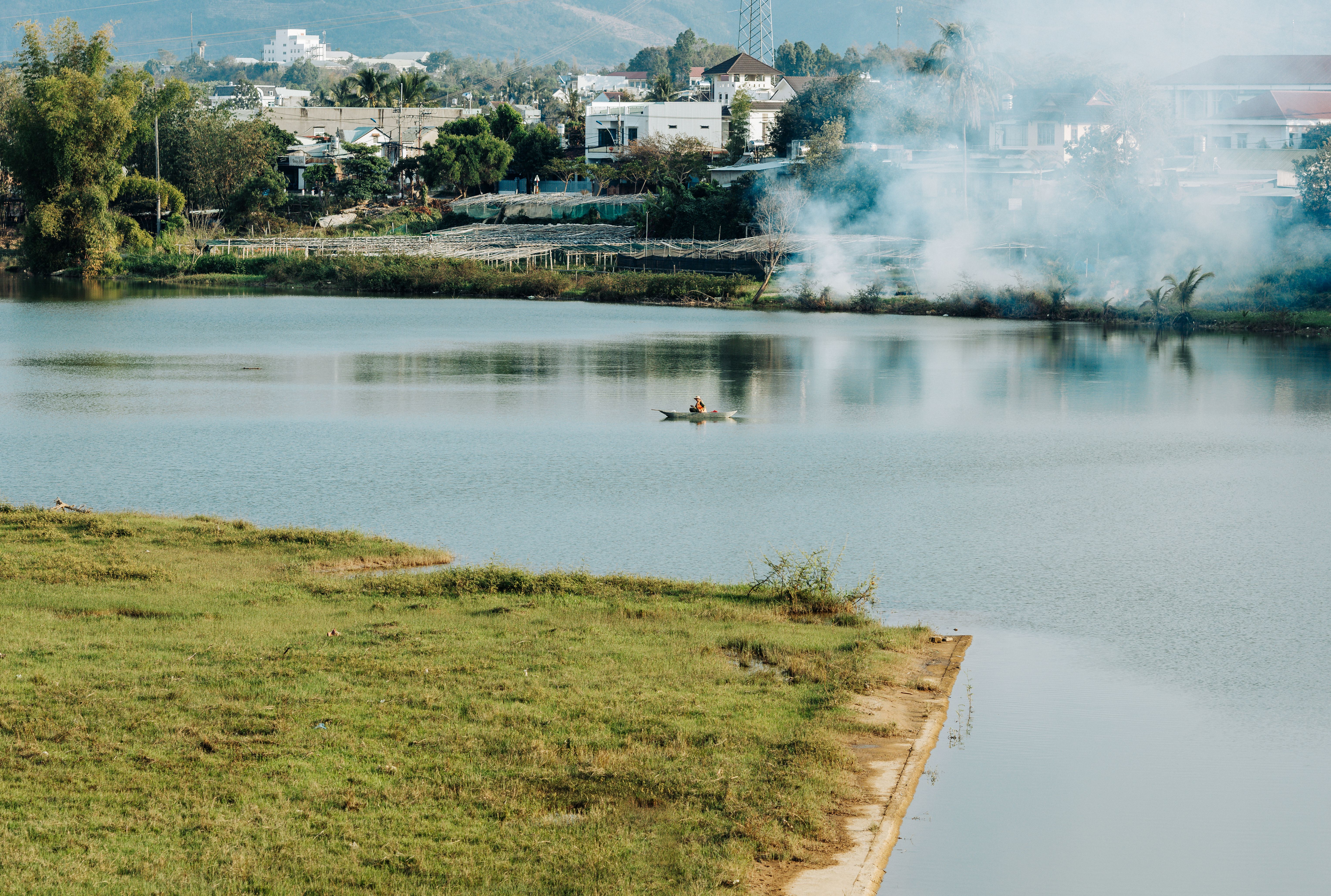 Peaceful lake view with lone canoeist in Kon Tum, Vietnam, framed by greenery and smoke in the distance.