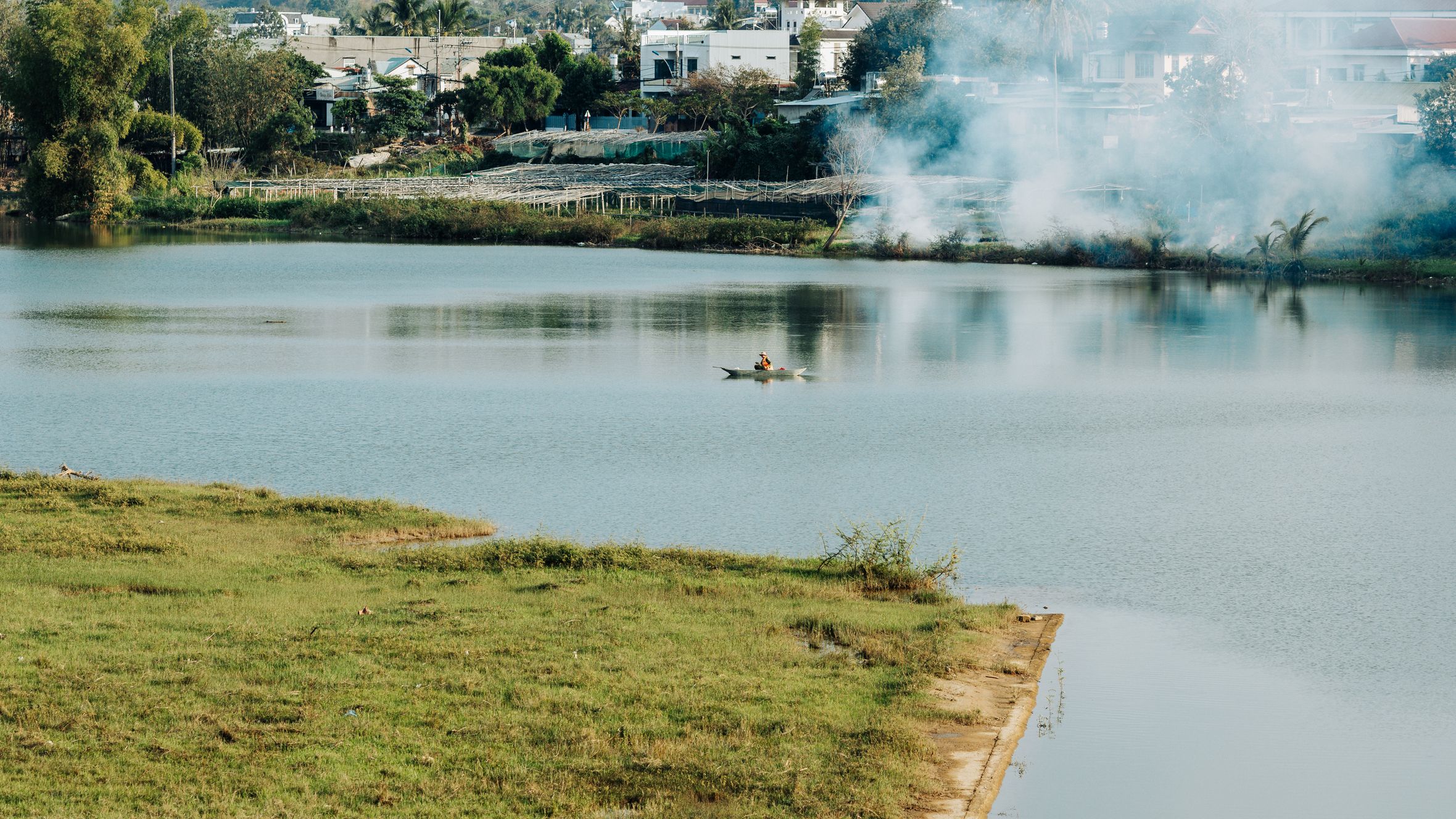 Peaceful lake view with lone canoeist in Kon Tum, Vietnam, framed by greenery and smoke in the distance.