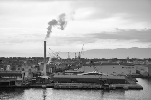 Black and white image of an industrial port with smoke rising from a factory stack and cranes in the background.