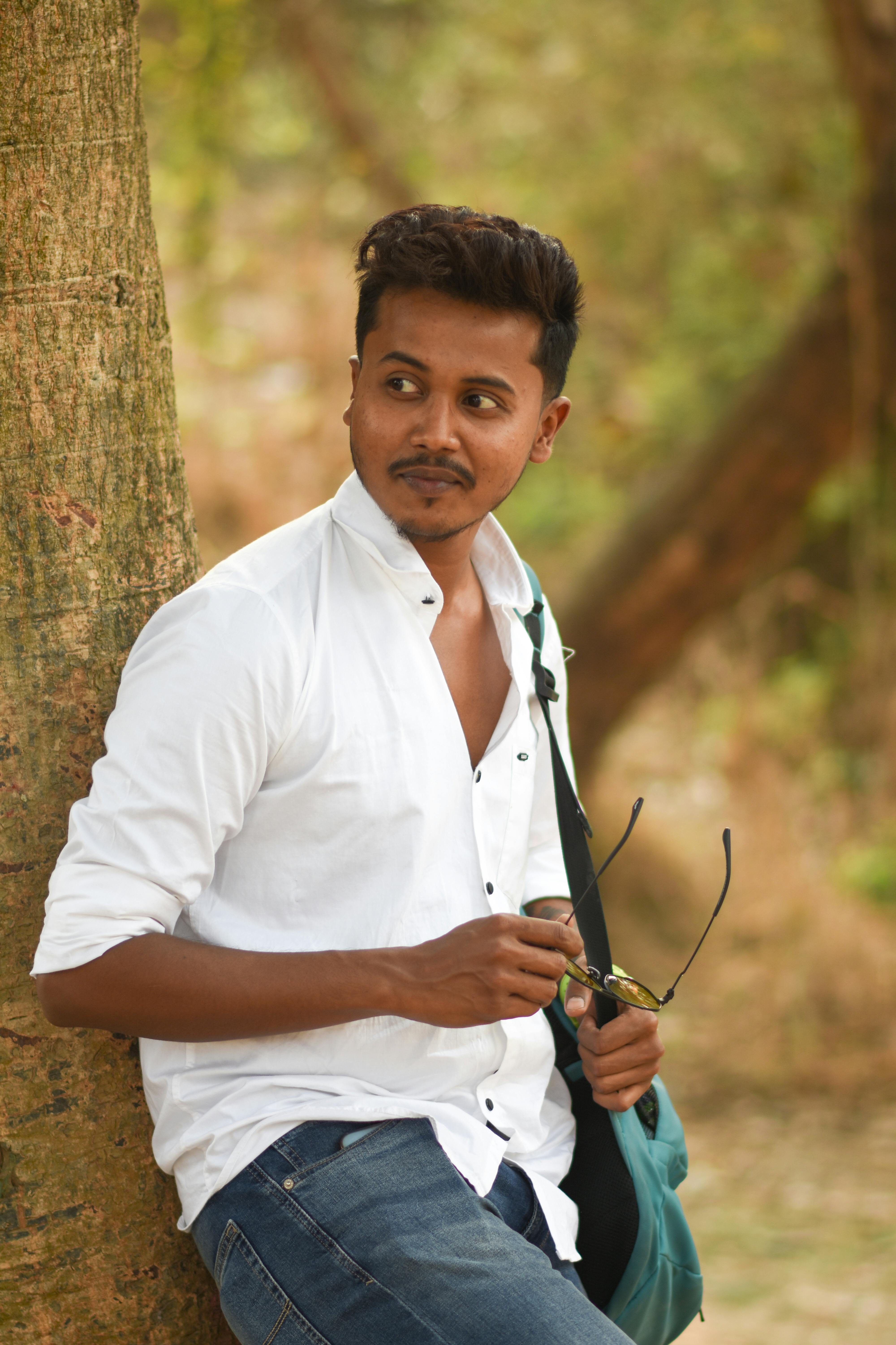 South Asian man in casual attire leaning on a tree, outdoors in Kolkata park, spring season.