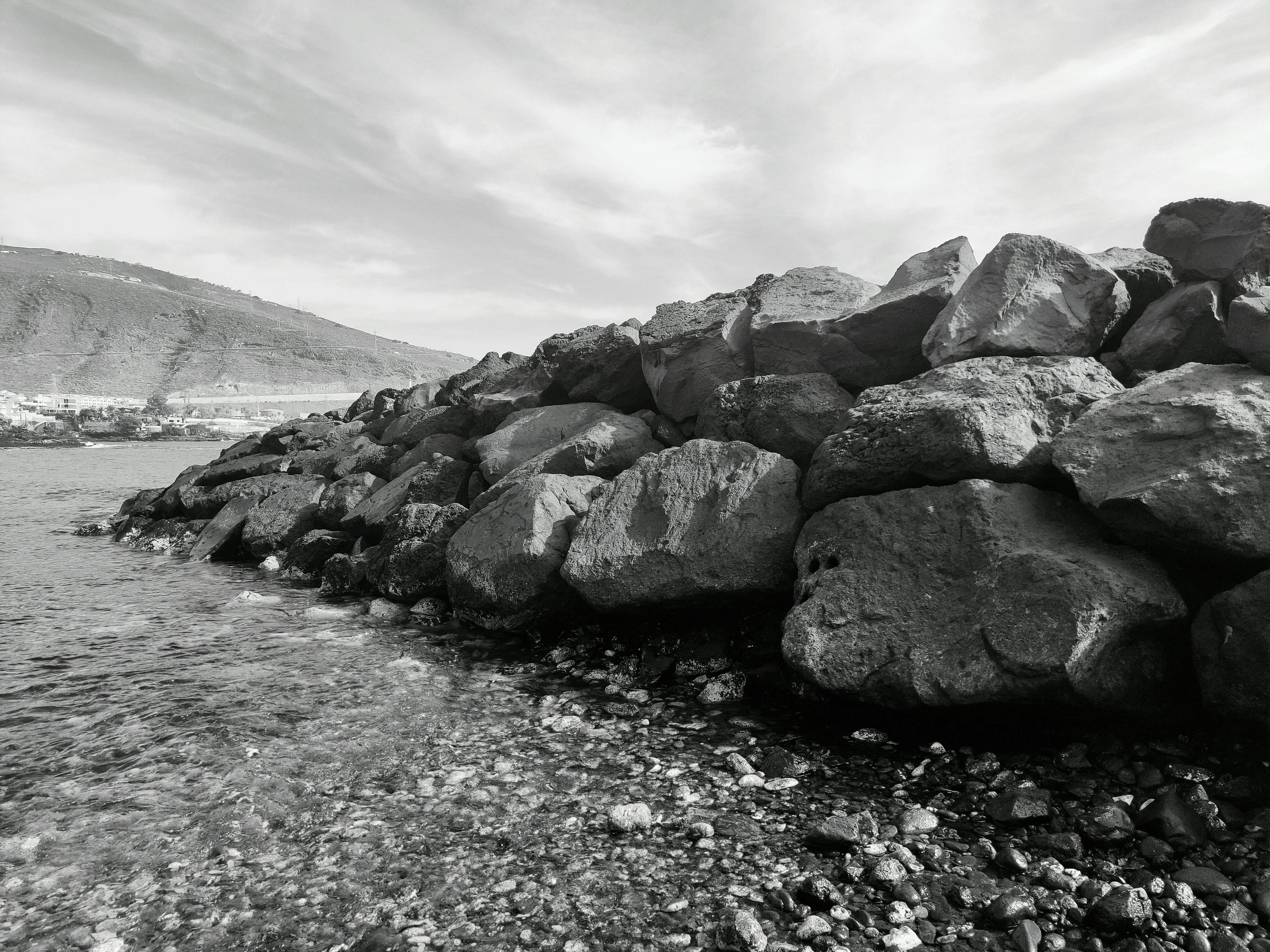 Gratis Et slående svart-hvitt-bilde av en steinete kystlinje som møter rolig vann under en overskyet himmel. Arkivbilde