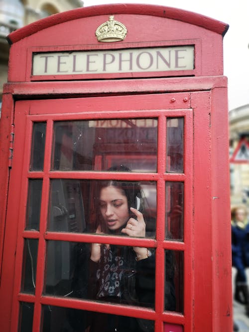 Woman in Black and White Floral Shirt Standing Inside Red Telephone Booth Making Call