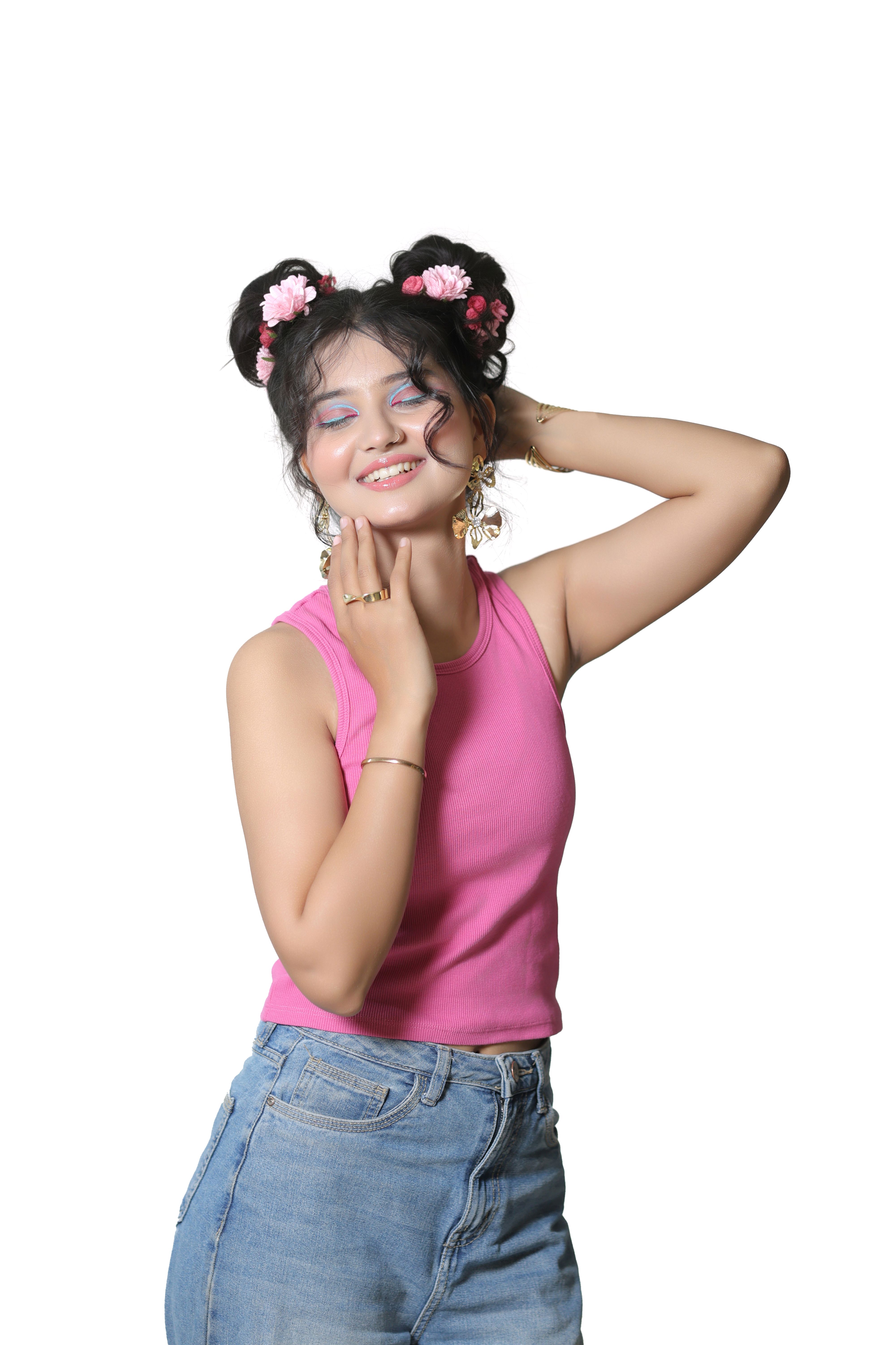 Smiling young woman with floral hair accessories, wearing pink top and denim, posing joyfully on a white background.