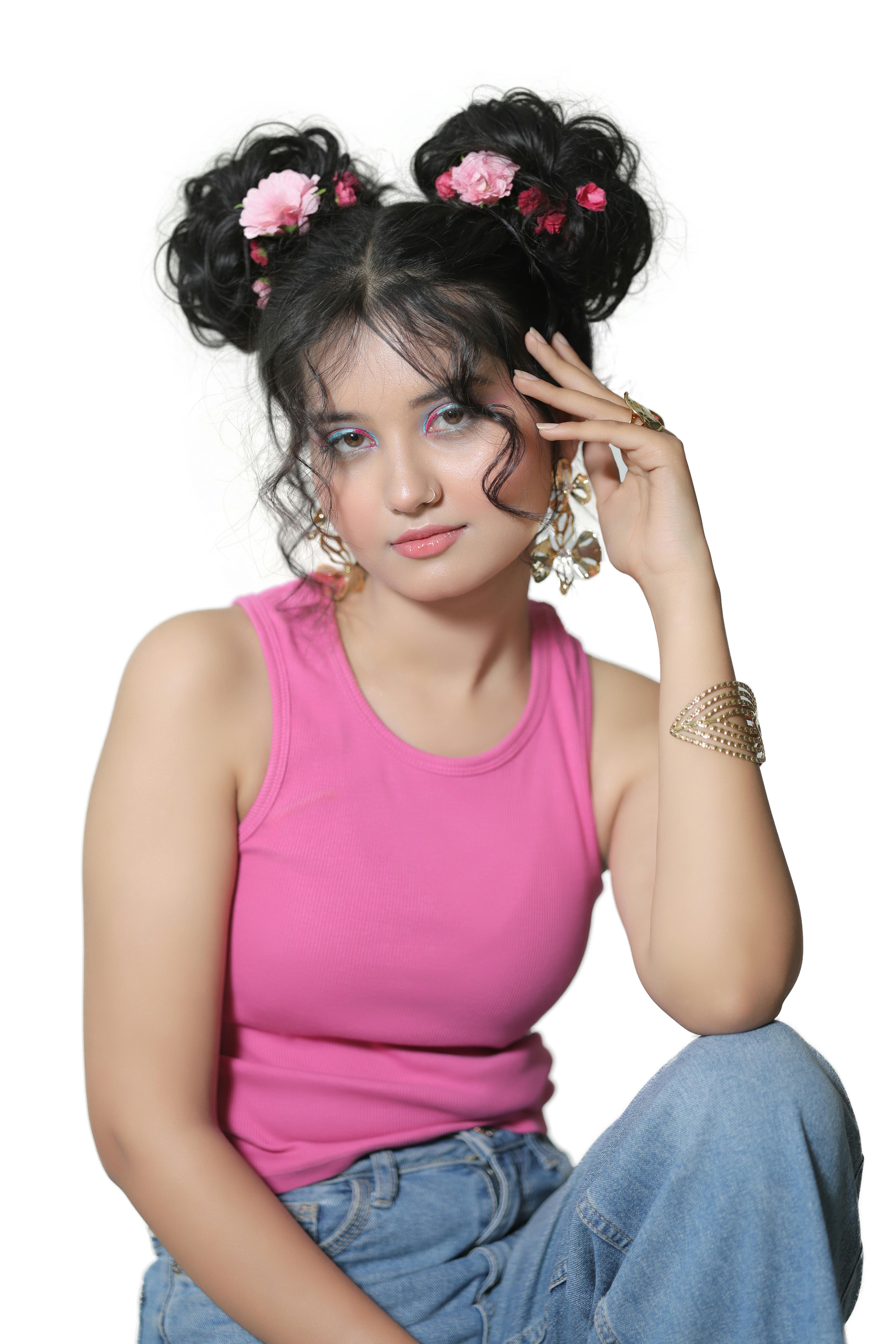 Free Stylish young woman with floral hair buns, wearing a pink tank top and denim, posing confidently indoors. Stock Photo