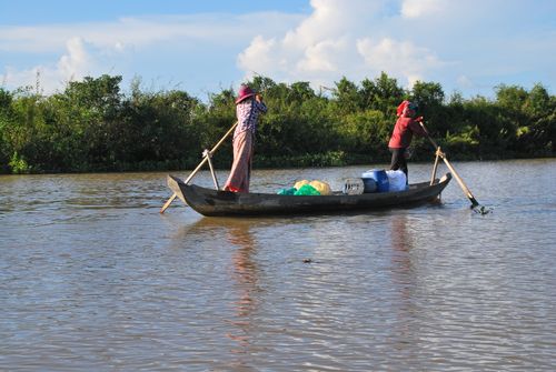 Free Two people maneuvering a traditional wooden boat on a calm river with greenery in the background. Stock Photo
