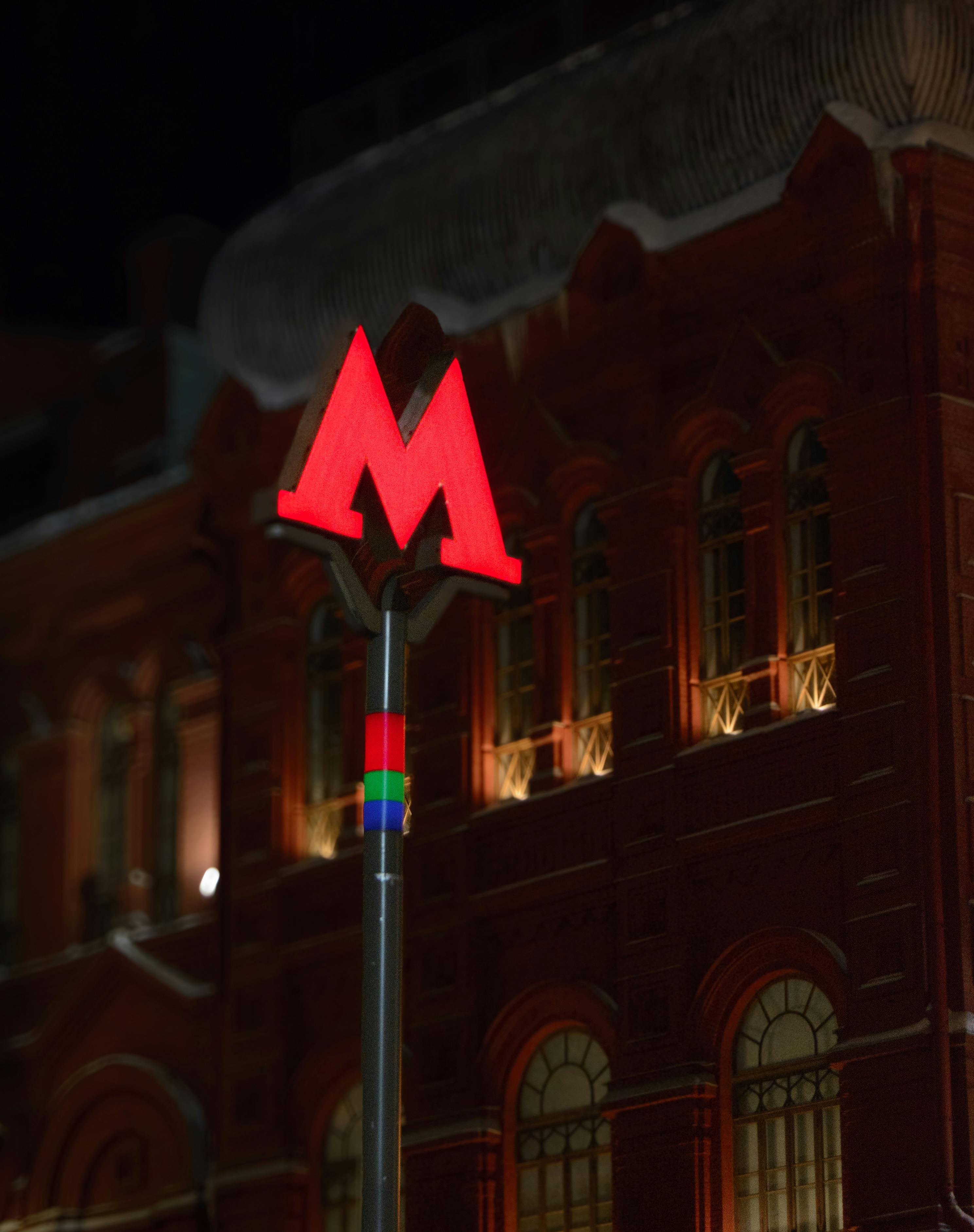 Gratis Rambu Metro Moskow berwarna merah yang menyala dengan latar belakang bangunan bersejarah di malam hari. Foto Stok