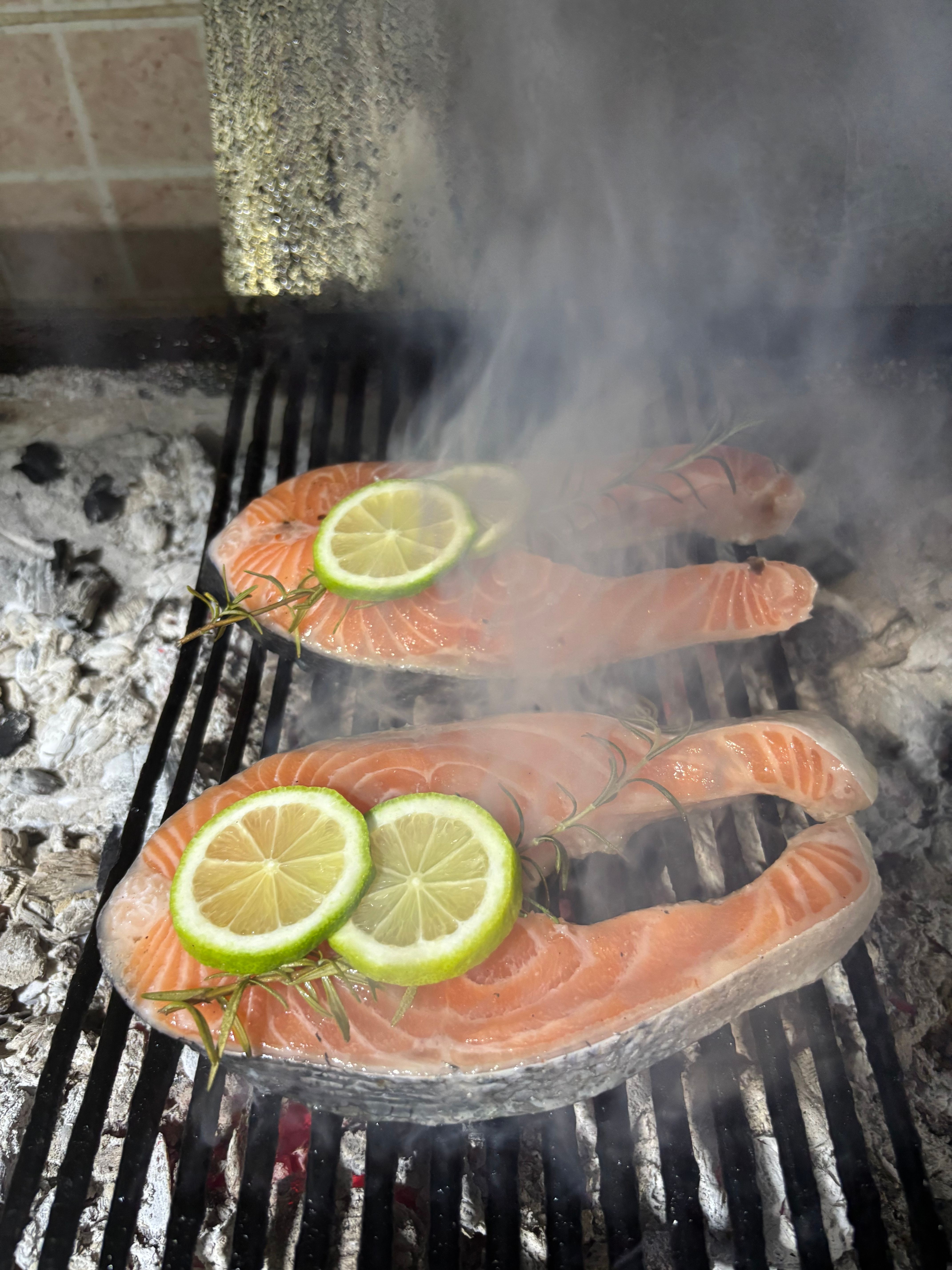Close-up of salmon steaks with lime slices and rosemary grilling over charcoal fire, emitting smoke.