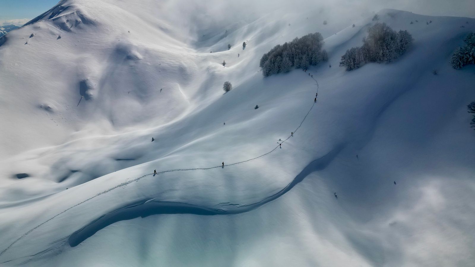 Aerial view of skiers climbing across snow-covered mountains under drifting cloud in Dibër County, eastern Albania