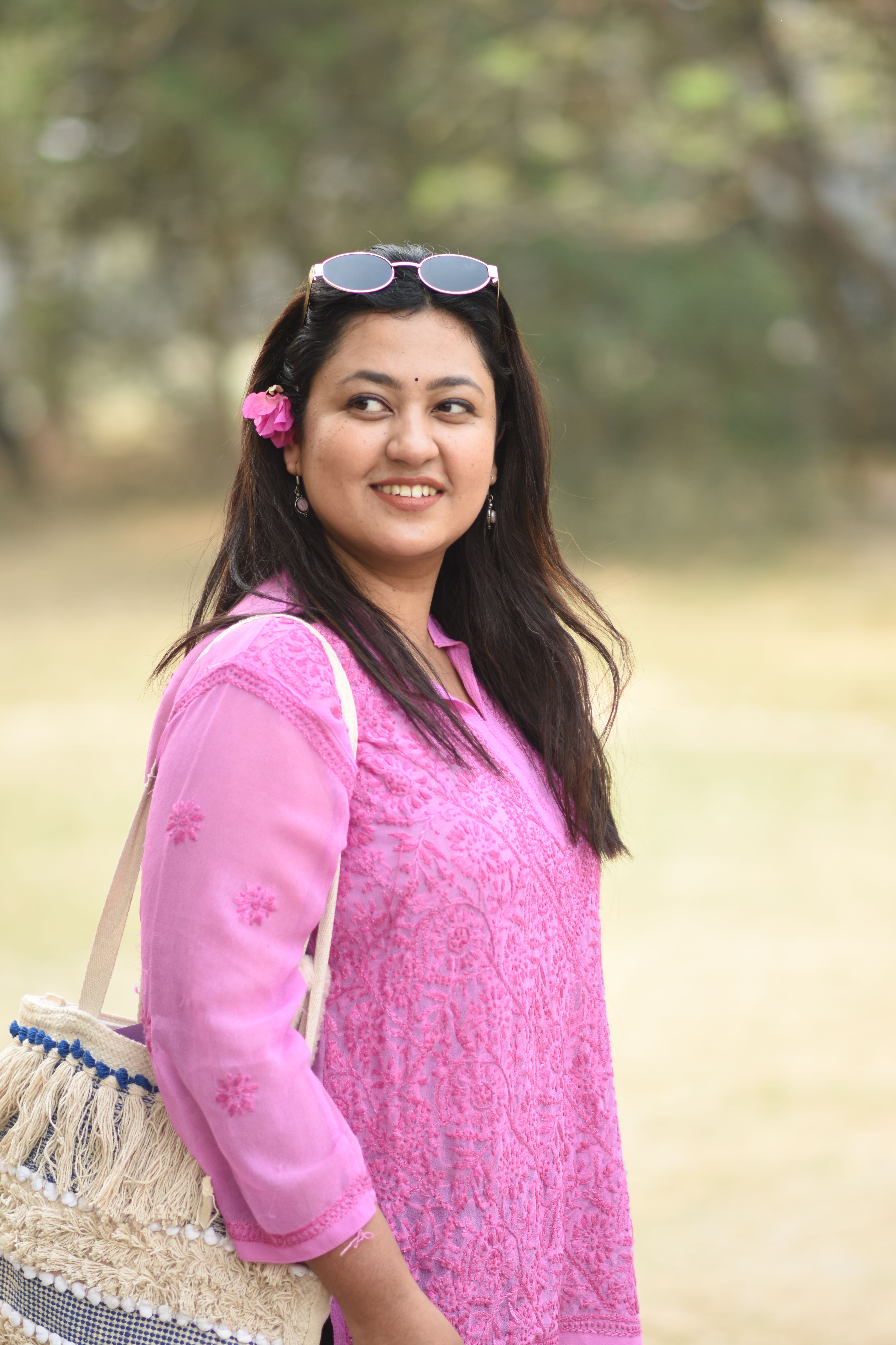 Portrait of a smiling woman in a pink dress outdoors with sunglasses and a bag.