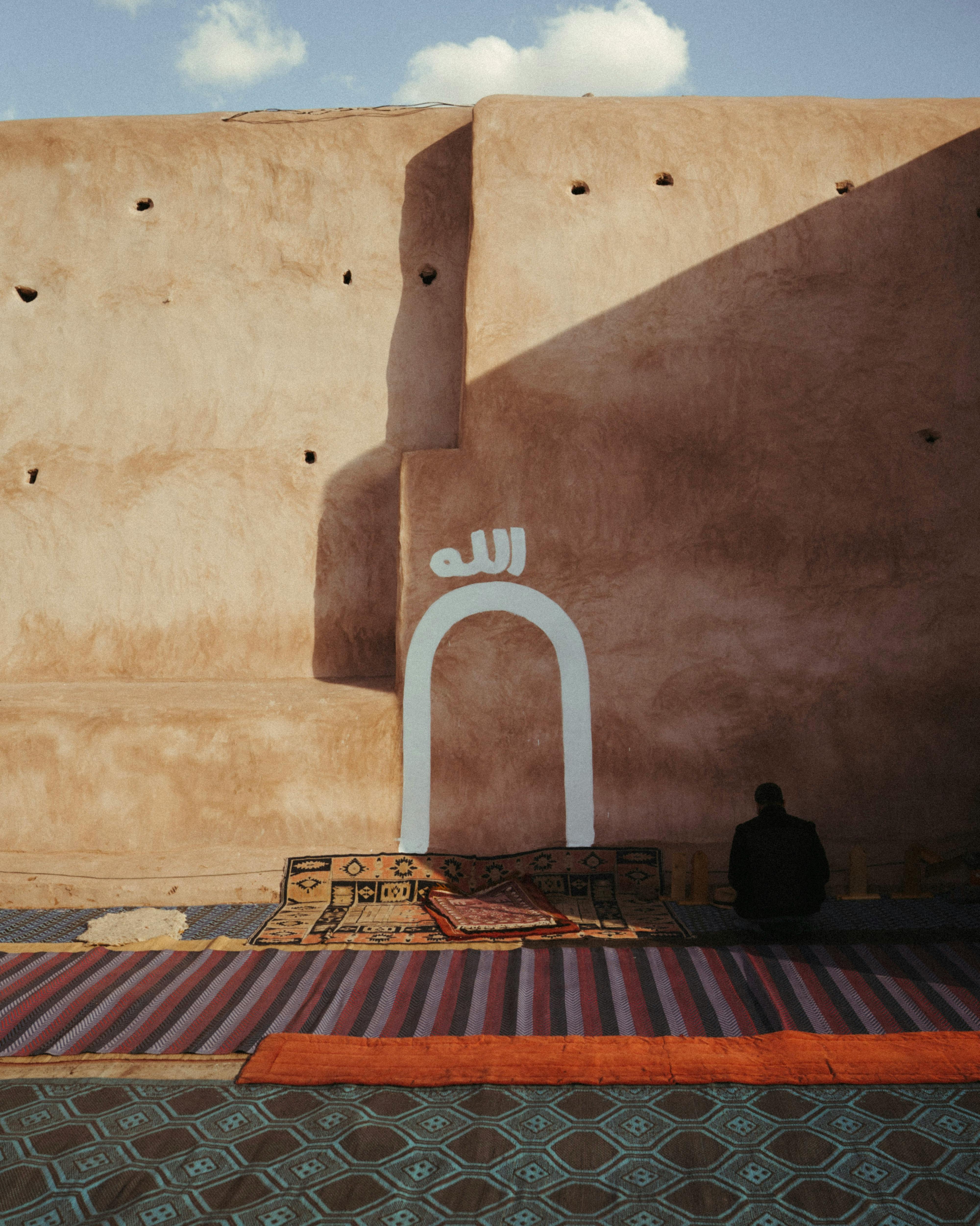 無料 モロッコのマラケシュにある、暖かい日差しと伝統的な絨毯に照らされた古代の壁のそばに男性が座っている。 写真素材