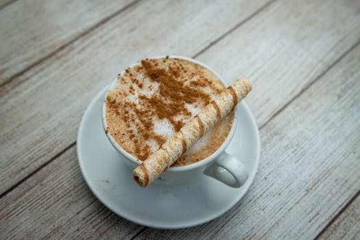 A cozy latte topped with cinnamon and a wafer stick served in a white cup on a wooden table.