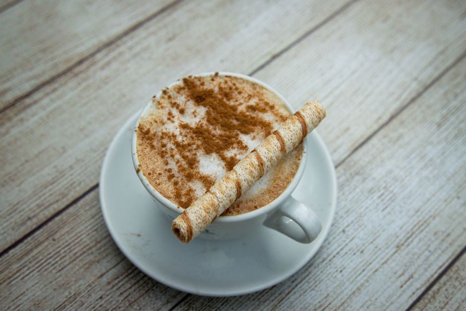 A cozy latte topped with cinnamon and a wafer stick served in a white cup on a wooden table.