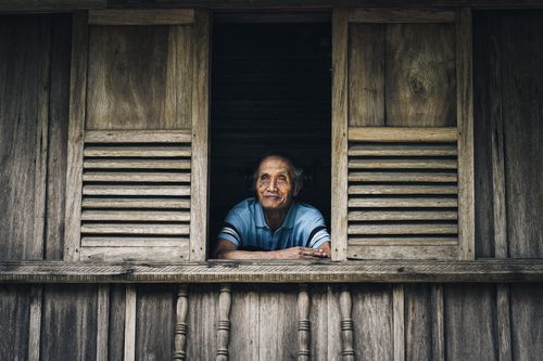 Free Elderly man in blue shirt gazing thoughtfully out a rustic wooden window, conveying nostalgia. Stock Photo