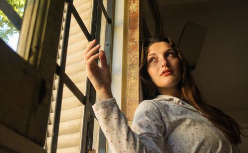 Free Young woman in floral shirt daydreaming by sunlit window indoors. Stock Photo