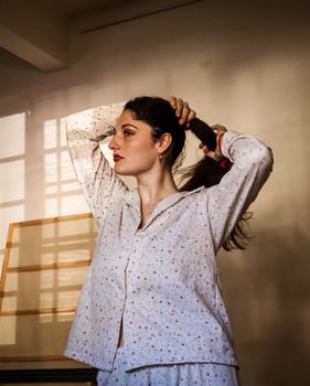 A young woman in pajamas stands indoors, styling her hair with natural sunlight streaming in.