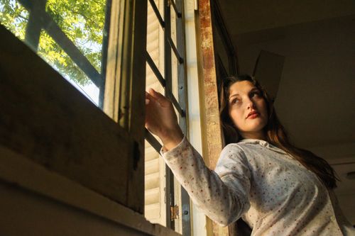Free A thoughtful woman stands by an open window, bathed in warm sunlight, indoors. Stock Photo