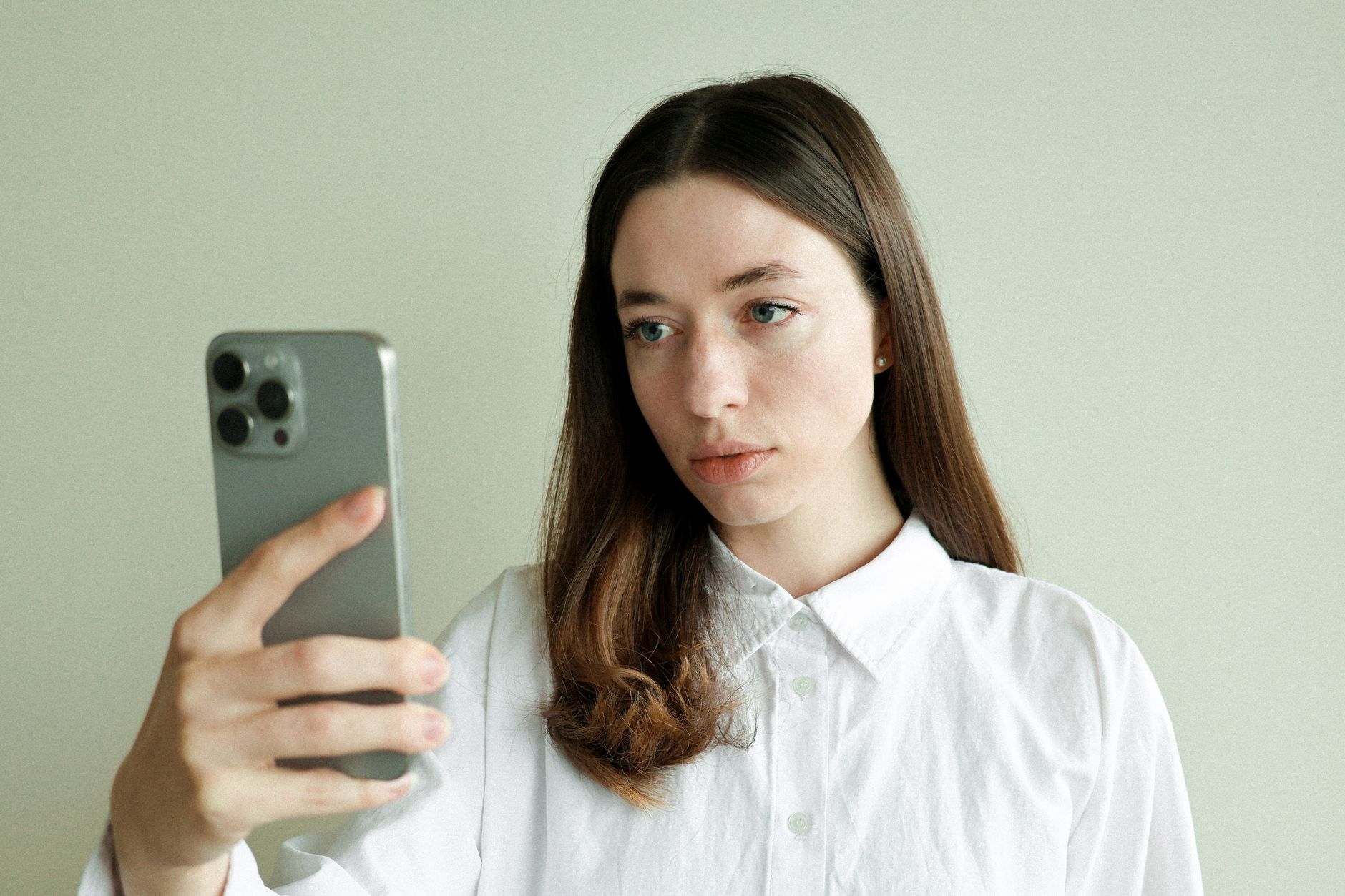Young woman with long hair takes selfie using smartphone indoors, neutral background.