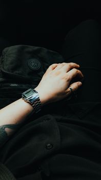 A stylish hand wearing a watch and rings with a visible tattoo, resting on dark clothing in low light.