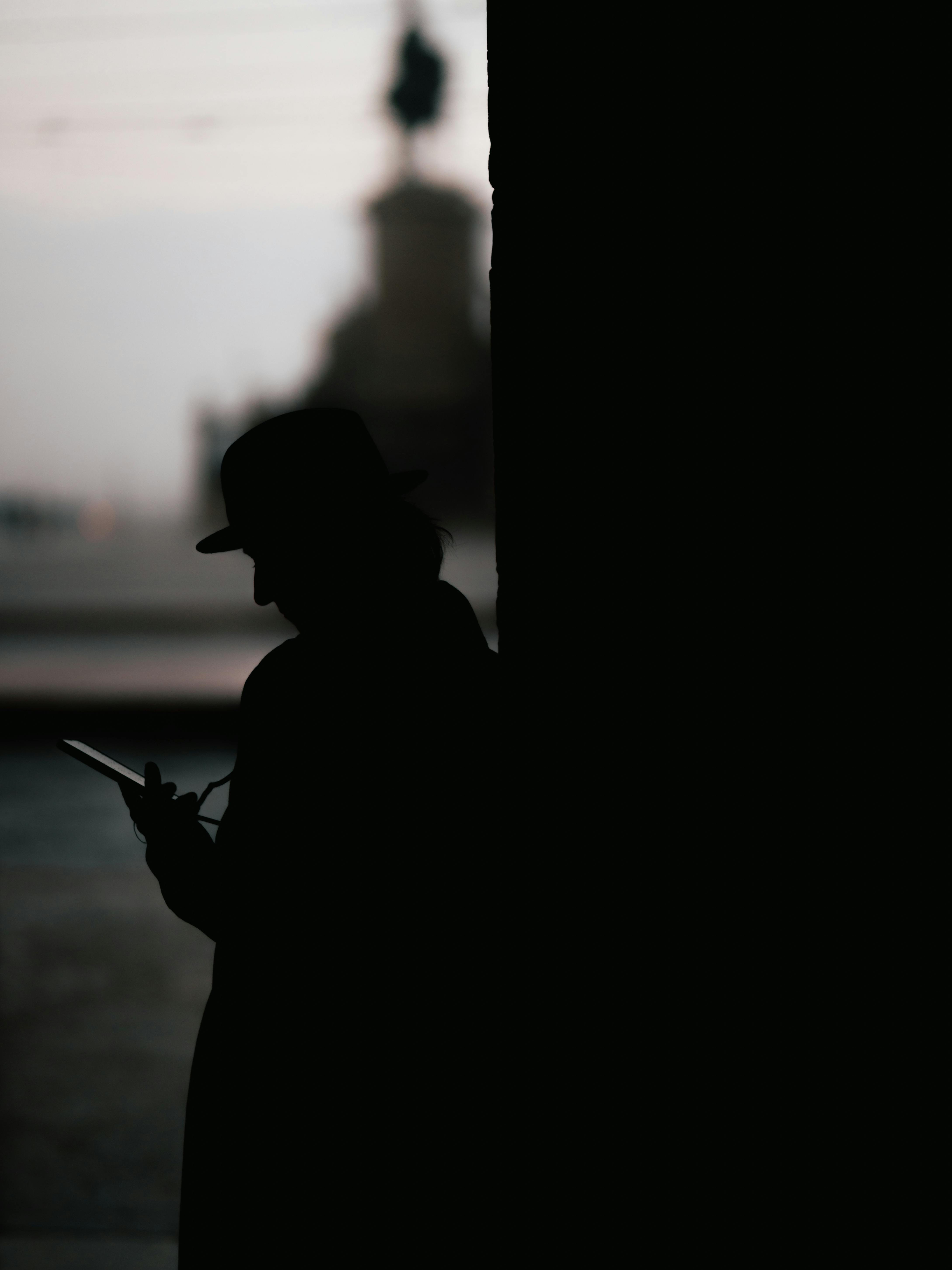 Δωρεάν Σιλουέτα ενός ατόμου με καπέλο που χρησιμοποιεί smartphone, απαθανατισμένη σε εξωτερικό χώρο με αστικό φόντο. Φωτογραφία από στοκ φωτογραφιών