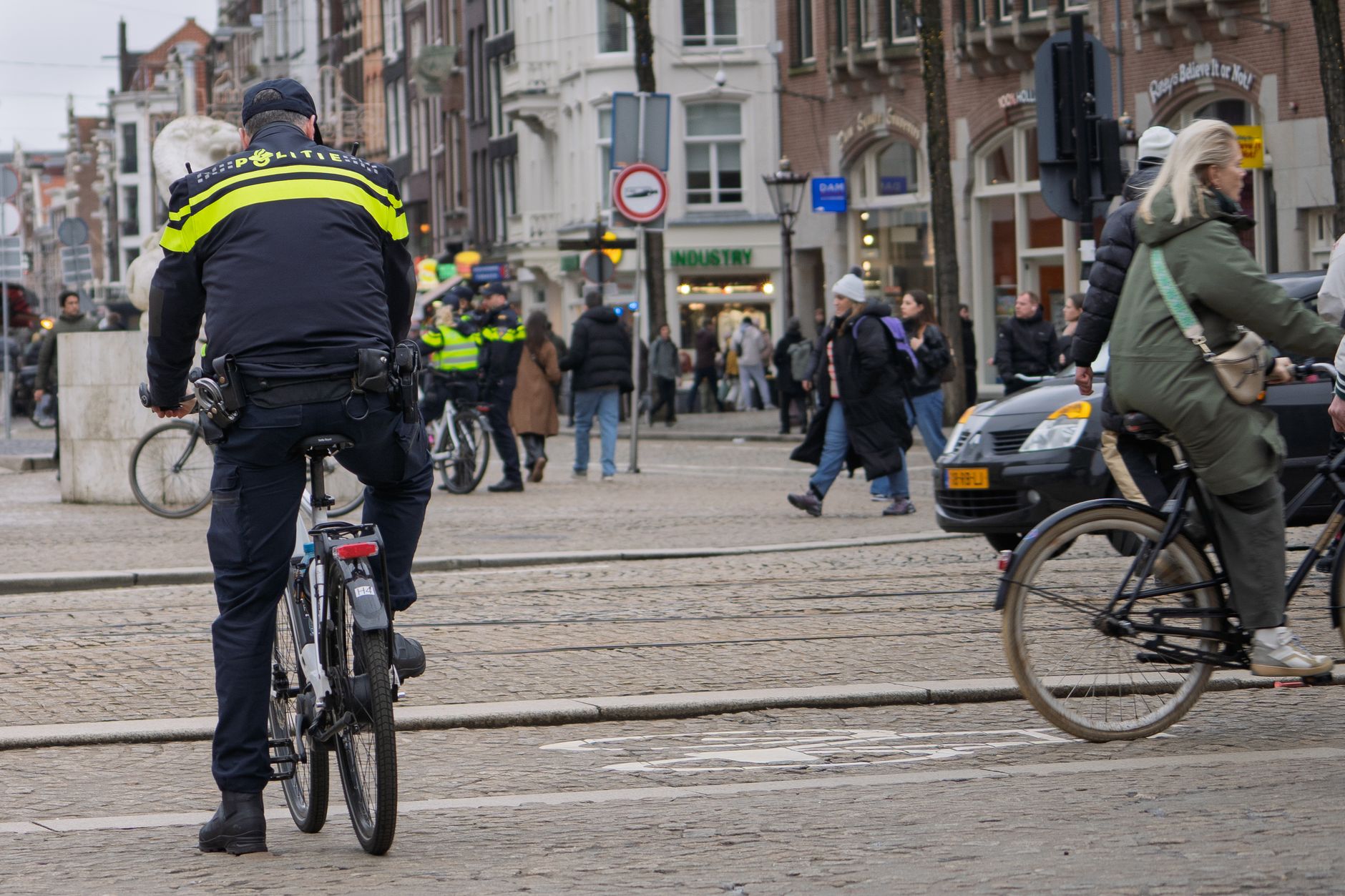 Een politieagent patrouilleert op een fiets door een drukke stadsstraat, met voetgangers en fietsers in de buurt.