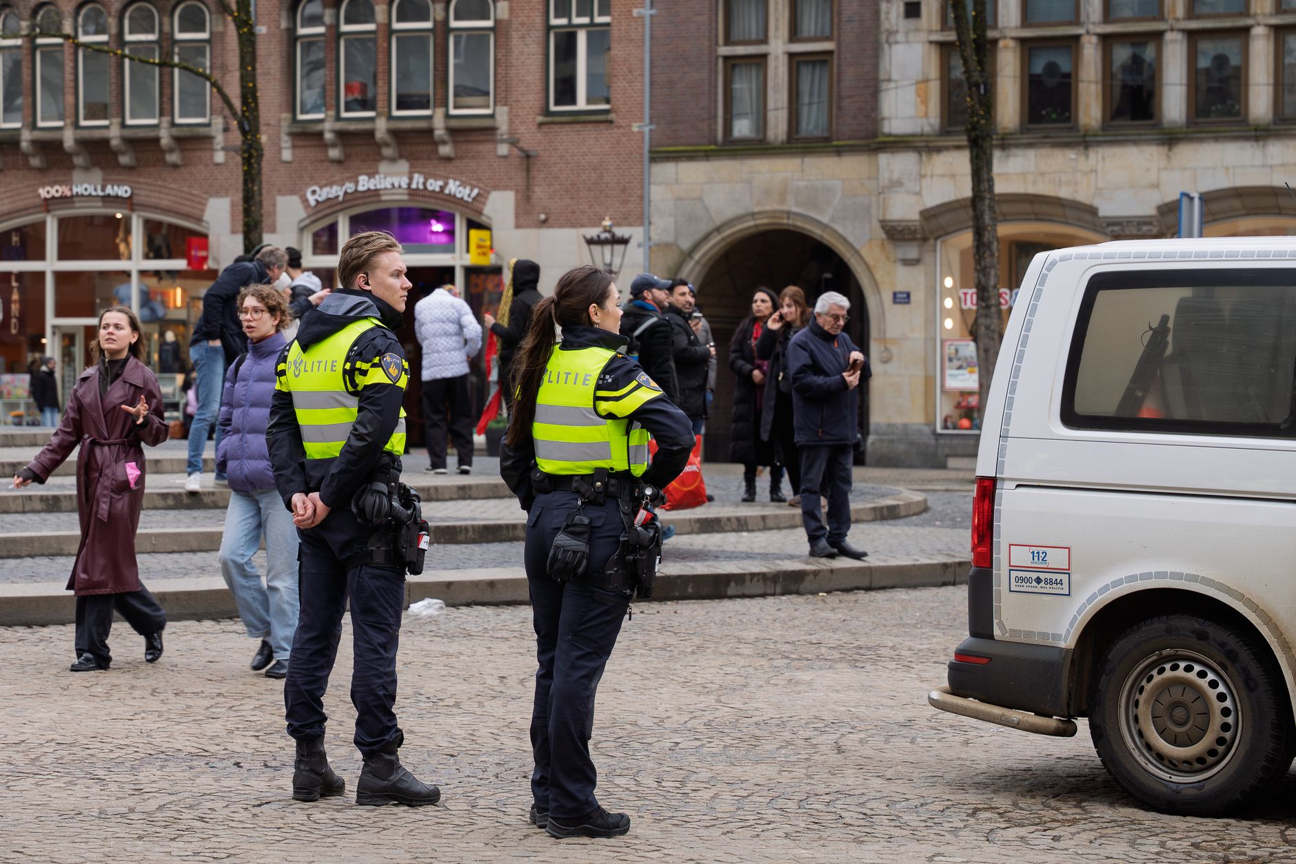 Stadsplein met Nederlandse politieagenten op patrouille, omringd door voetgangers en een politieauto.