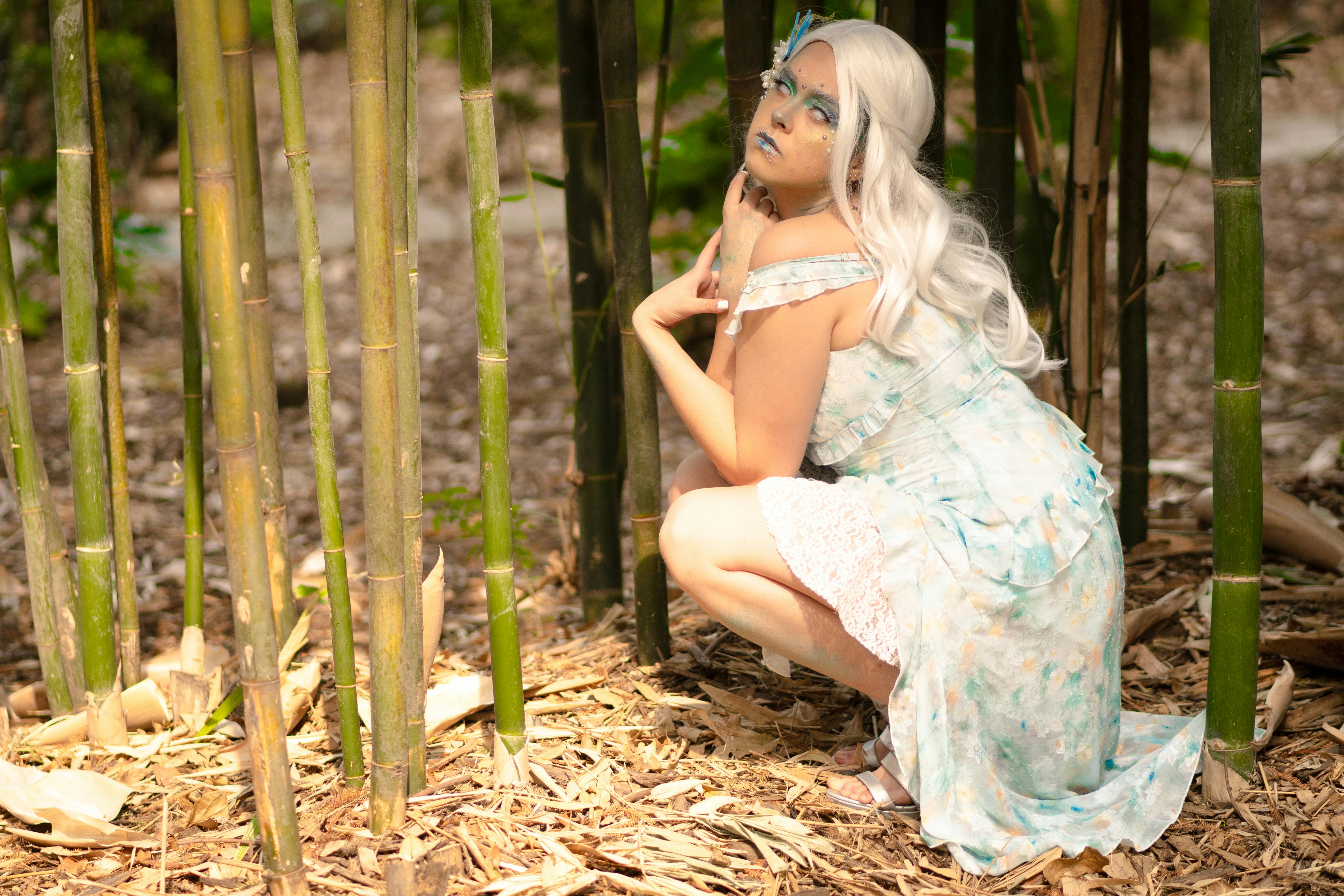 Gratis Seorang wanita dengan riasan artistik berjongkok di antara bambu, mengenakan kostum bertema fantasi. Foto Stok