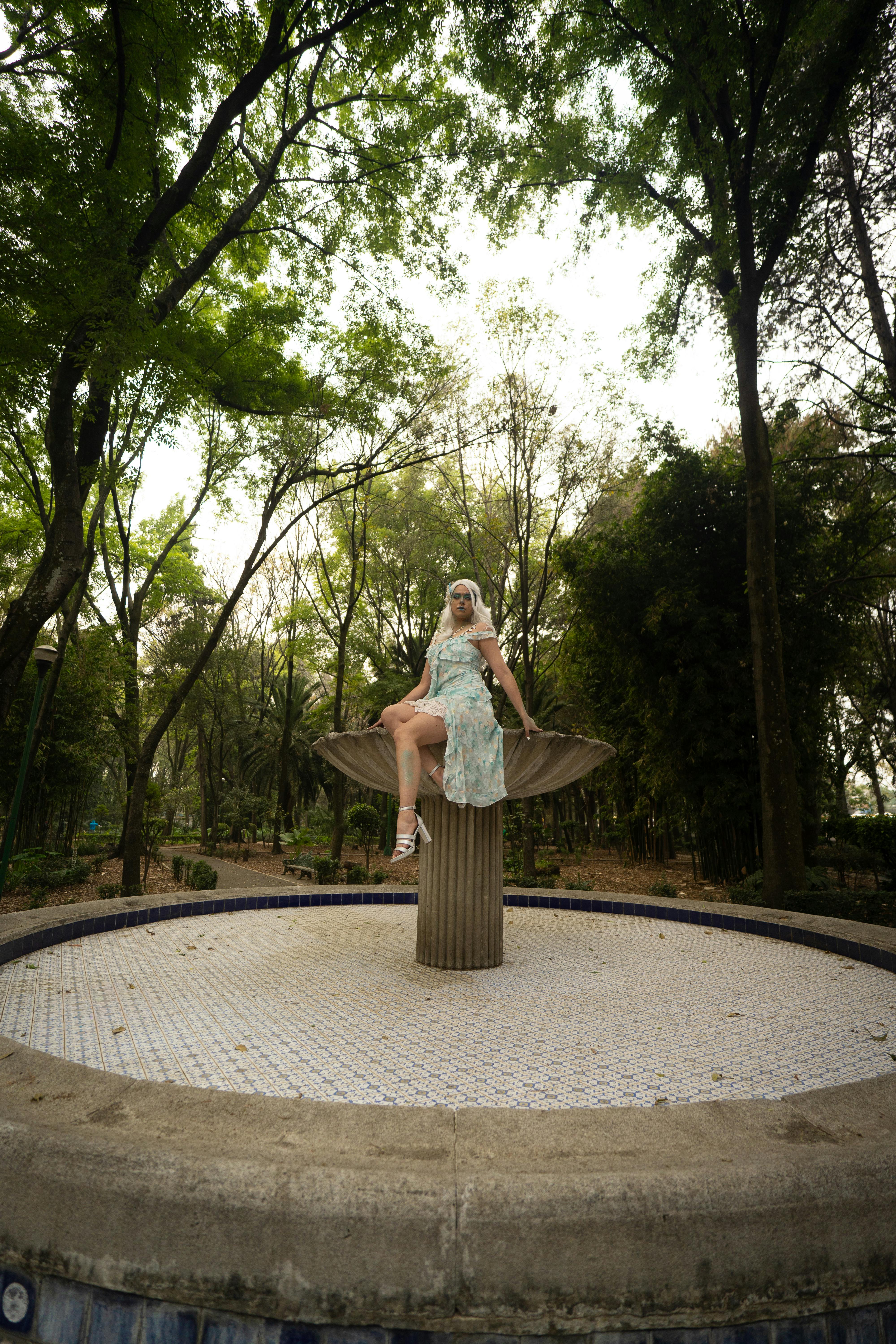 бесплатная Молодая латиноамериканка в художественном костюме позирует у фонтана в живописном общественном парке. Стоковое фото